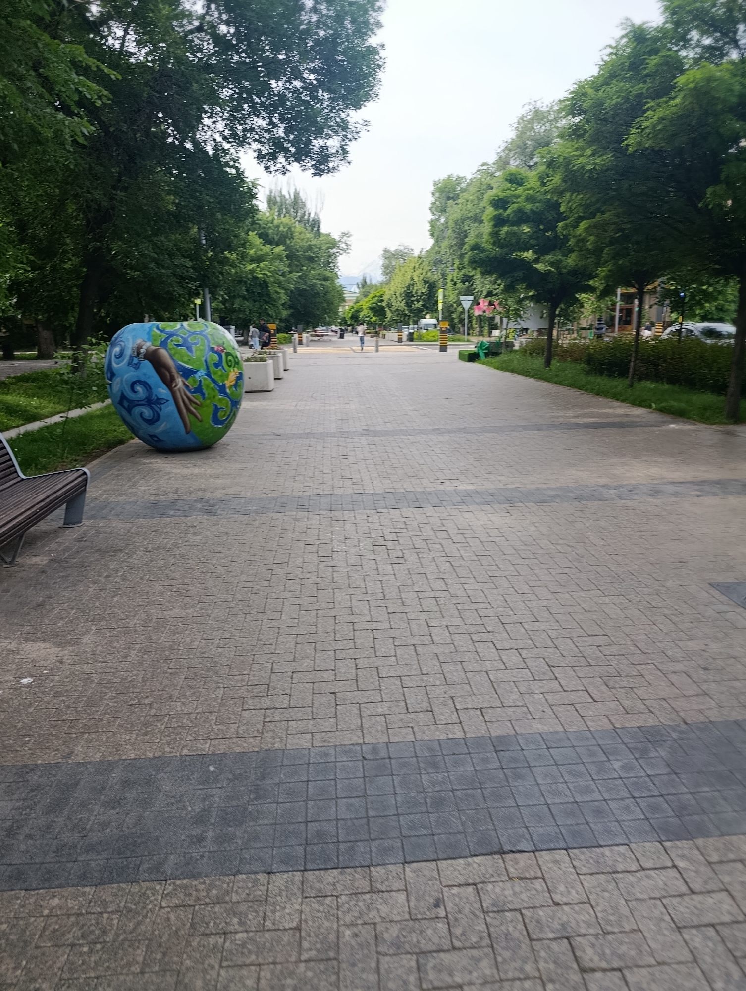 The image depicts a wide, paved pedestrian walkway in Almaty, Kazakhstan. The walkway is constructed of light gray rectangular pavers with a dark gray paver strip running horizontally across the foreground. The walkway is flanked by green trees and grass on both sides. On the left side of the walkway is a large, spherical sculpture resembling an apple, painted with blue and green designs and a hand. A wooden bench with metal legs is also visible on the left, partially obscured by the sculpture. Further down the walkway, several people are walking in the distance, and there are bollards and planters. In the background, buildings and distant mountains can be seen. The sky is overcast.