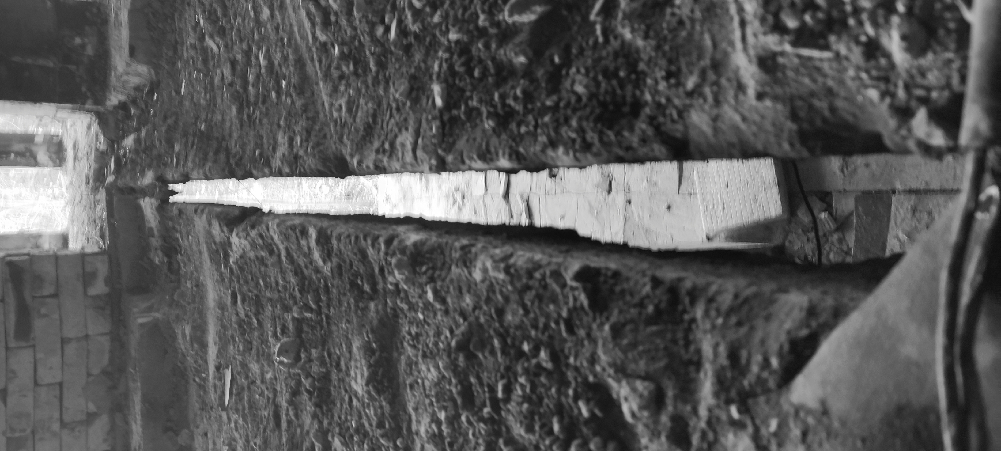 The image is a black and white, close-up shot of what appears to be the interior of a dilapidated structure, possibly located in Fryazino, Russia. The central focus is a narrow horizontal gap or opening between two roughly textured, possibly concrete or stone surfaces. Through this opening, a glimpse of a brighter area is visible, showing fragmented views of architectural elements like walls and beams. To the left of the gap, there's a section of brickwork, possibly part of a wall. The overall impression is one of decay and disrepair. The lighting is uneven, creating strong contrasts and shadows that emphasize the textures of the surfaces. There are no discernible people or distinct actions taking place in the image. The environment appears to be abandoned or under construction.