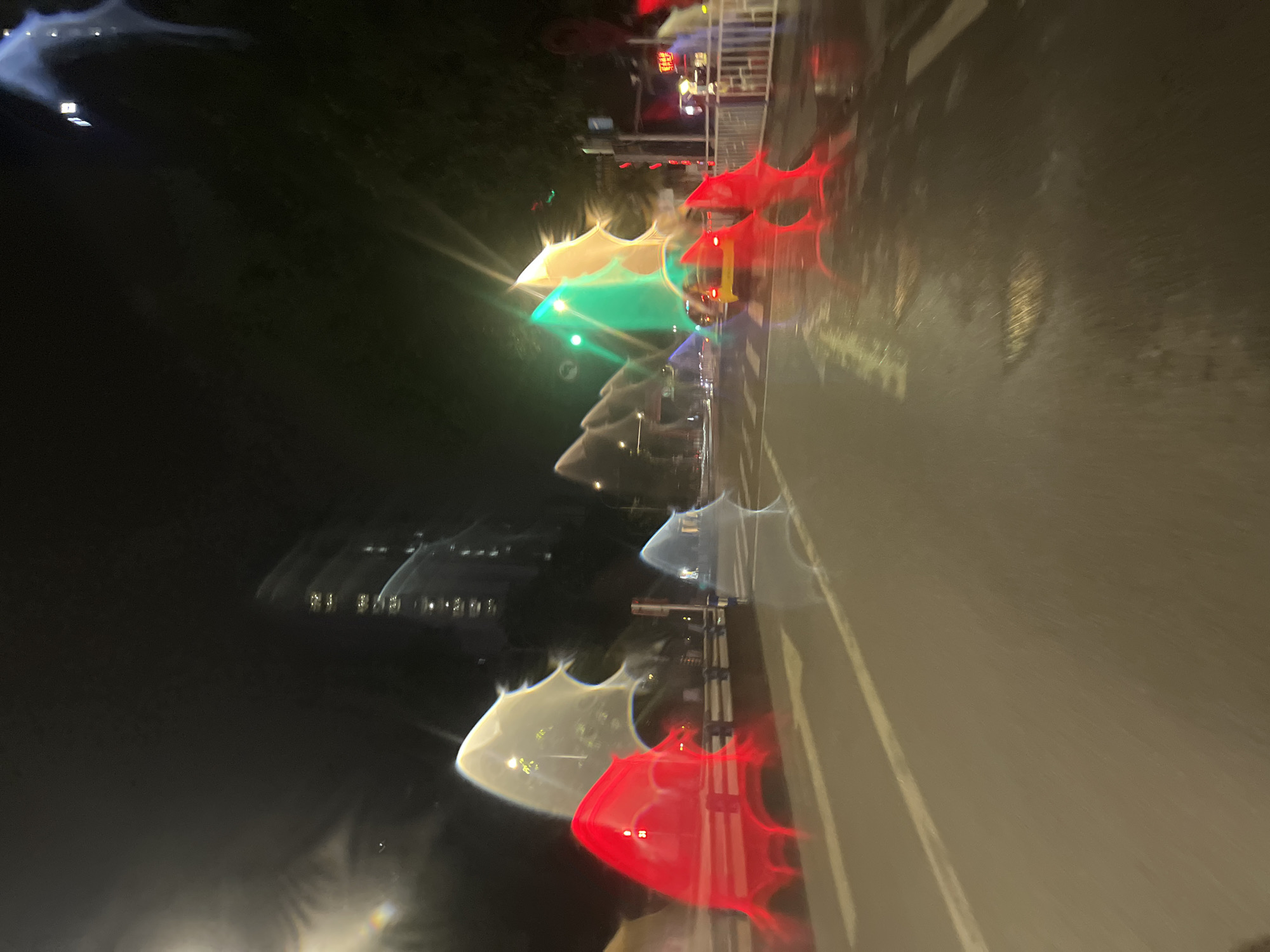 The image depicts a nighttime scene on a wet road, possibly a city street, in Jiulongpo District, China. The perspective appears to be from inside a vehicle, looking forward. Dominating the foreground are abstract, distorted red and white light refractions, suggesting they are reflections or light sources viewed through a wet surface like a windshield. In the mid-ground, a yellow car is visible, also with distorted lights. The road surface is wet and reflective, with lane markings visible. Beyond the road, buildings are present, some illuminated with lights, and trees line the side of the street. Traffic lights are also discernible, with at least one green light illuminated. The overall impression is a blurry, somewhat surreal urban nightscape due to the optical effects and the wet conditions.