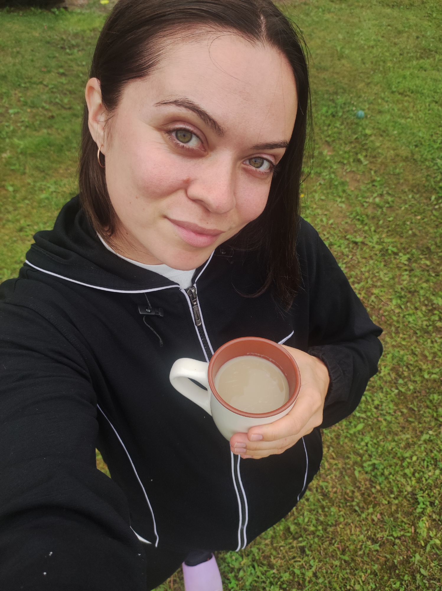 A woman is taking a selfie outdoors, holding a mug of light brown liquid. She has dark brown hair, light skin, and green eyes. She is wearing a black hooded jacket with white trim and a white t-shirt. Her left hand is holding the mug, which has a beige exterior and a brown rim. The background is a grassy area. The specific location in the city of Фоки, Russia, is not discernible from the image.