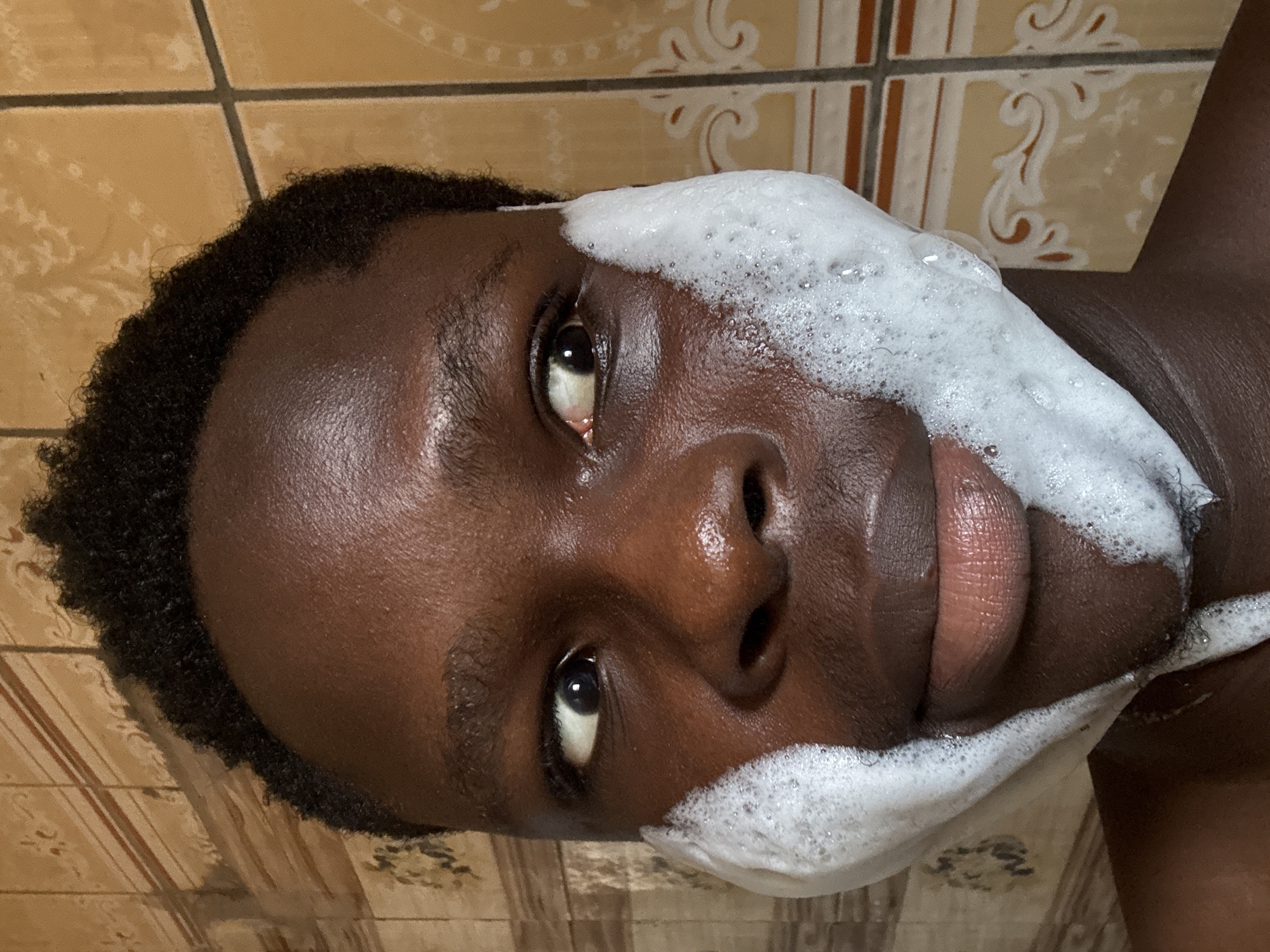 A close-up, vertically oriented photo shows a person's face, with a portion of their head and upper body visible. The person has dark skin and dark, curly hair. Their face is partially covered in white foam, suggesting they are washing their face or hair. The foam is concentrated on the right side of their face and extends towards their forehead and cheek.
The person's eyes are open and looking towards the camera. Their expression is neutral. The background consists of tiled walls, with the tiles appearing to be a light brown or beige color with darker grout lines. Some of the tiles have a decorative pattern.
The lighting is bright, indicating it's likely daytime. The setting appears to be indoors, possibly a bathroom or shower area, given the presence of tiles and foam. The location context is Ihiagwa, Nigeria. There is no visible text in the image.