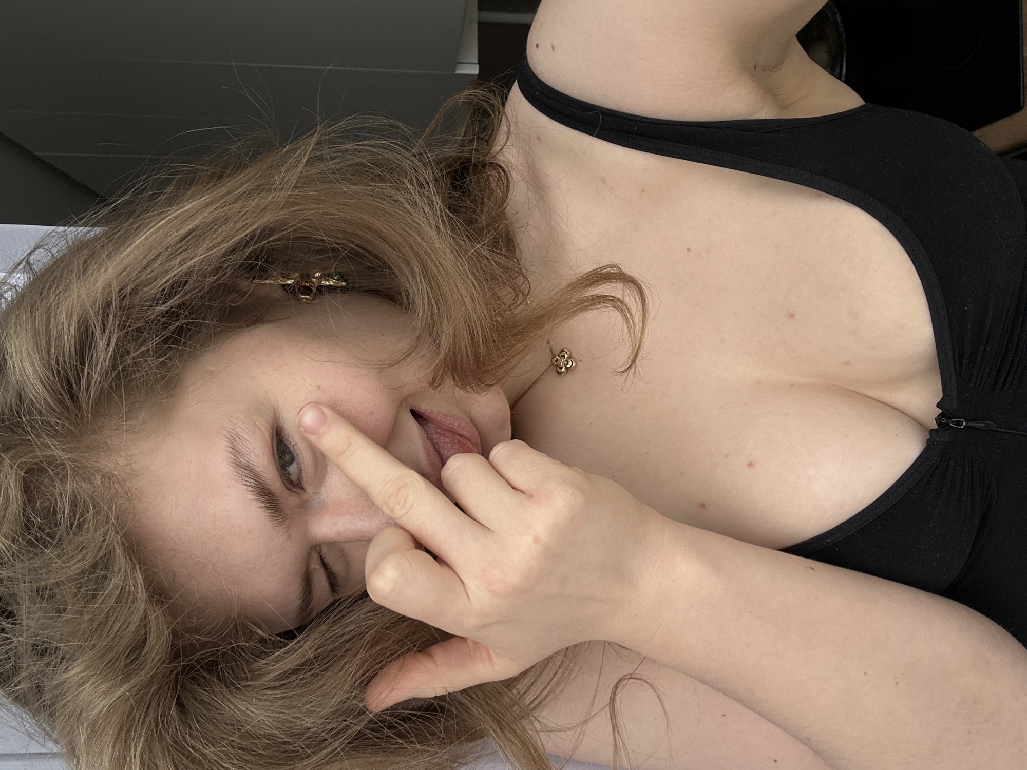 A woman with light-brown hair is lying down, wearing a black top. Her left hand is raised, obscuring part of her face, and her tongue is slightly visible. She has an earring on her left ear and a necklace. The background is out of focus. There's no information to suggest the photo was taken in Kazan, Russia.