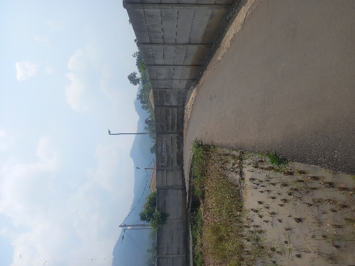 The image depicts a rural or semi-urban landscape in Mekarjaya, Indonesia, under a partly cloudy sky. A paved asphalt road, dark grey in color, curves gently from the lower center towards the upper right. To the left of the road is a water-logged field or ditch containing numerous short, light brown stubble, possibly remnants of harvested crops, submerged in shallow, muddy water. Green weeds and grass grow along the immediate edge of the road and within the field.
A tall, segmented concrete wall, light grey and showing signs of weathering, runs along the right side of the road, mirroring its curve. Another section of a similar concrete wall is visible beyond the water-logged field on the left. Behind the walls, various objects are visible: on the left, a utility pole with power lines and a streetlamp; further right, a taller, slender streetlamp. Several low-rise buildings with light-colored roofs are partially obscured by the walls and sparse green foliage. In the distant background, multiple layers of hazy blue-grey mountains rise under a pale blue sky dotted with scattered white clouds. No individuals or active subjects are present.