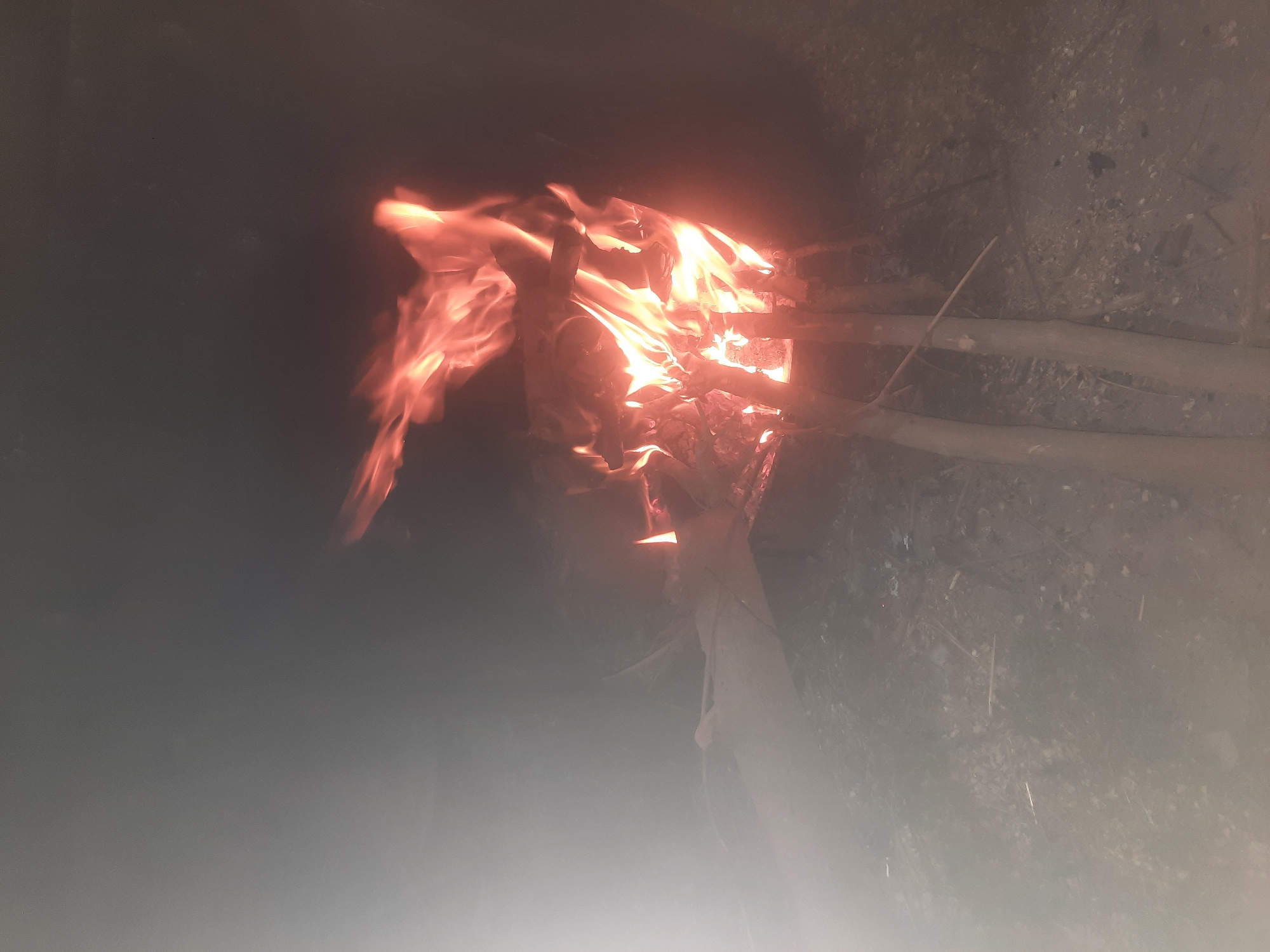 A bonfire burns in a dark, outdoor environment. The fire is contained within a makeshift structure of logs, with flames leaping upwards and casting an orange glow. Smoke billows around the fire, particularly on the left side of the image, creating a hazy effect. The ground beneath appears to be composed of dirt and debris. The scene is dimly lit, suggesting it is nighttime. No people are visible in the image. The location is identified as Voseaabad, Tajikistan.