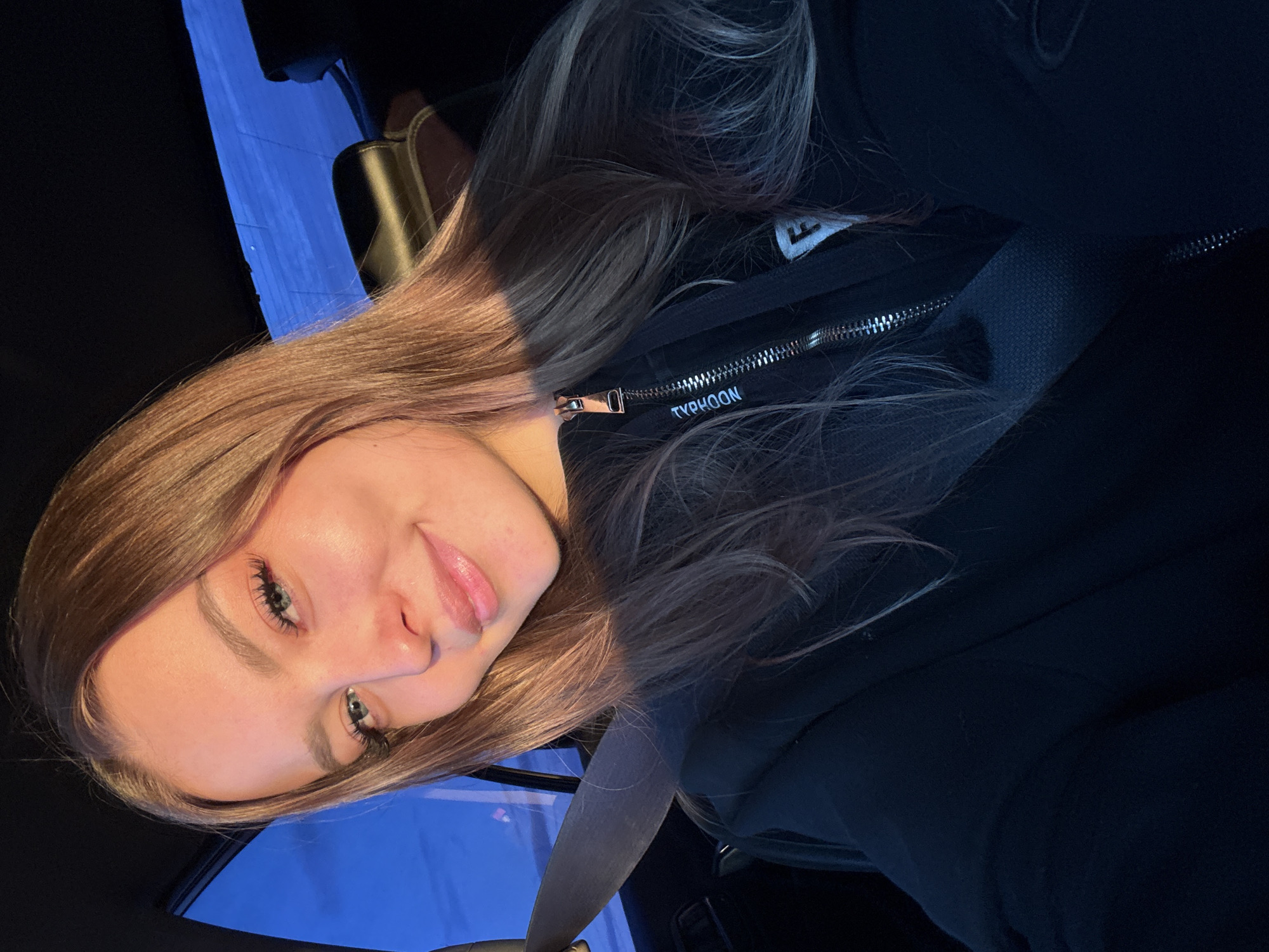 A woman is posing for a selfie while inside a car. She is wearing a black hoodie, with the word "TYPHOON" visible on the zipper area. A seatbelt is positioned across her chest. The light suggests it might be dusk or dawn. The location of the city of Tulagino, Russia, is not discernible from the image.