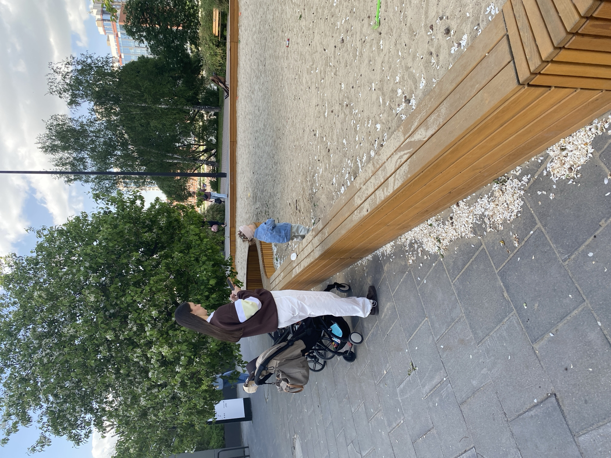 The scene depicts a partially obstructed view of a park-like environment, likely in an urban setting. In the foreground, a young woman with long, dark hair, wearing a brown sweater and white pants, stands next to a baby stroller. She appears to be looking at her mobile phone.
The stroller is light brown in color. The woman is standing on a paved surface constructed of gray rectangular stones. The surface is littered with scattered white flower petals.
Adjacent to the paved area is a light-colored wooden bench. Further beyond the bench is a sand-filled area.
In the background, trees and a partially visible building can be observed. Based on the visual cues provided, and the prompt request for Yekaterinburg, Russia, it is plausible this could be a park in Yekaterinburg, though specific landmarks are not discernible to pinpoint the exact location within the city.
Other individuals are visible in the background, including a child and several adults.