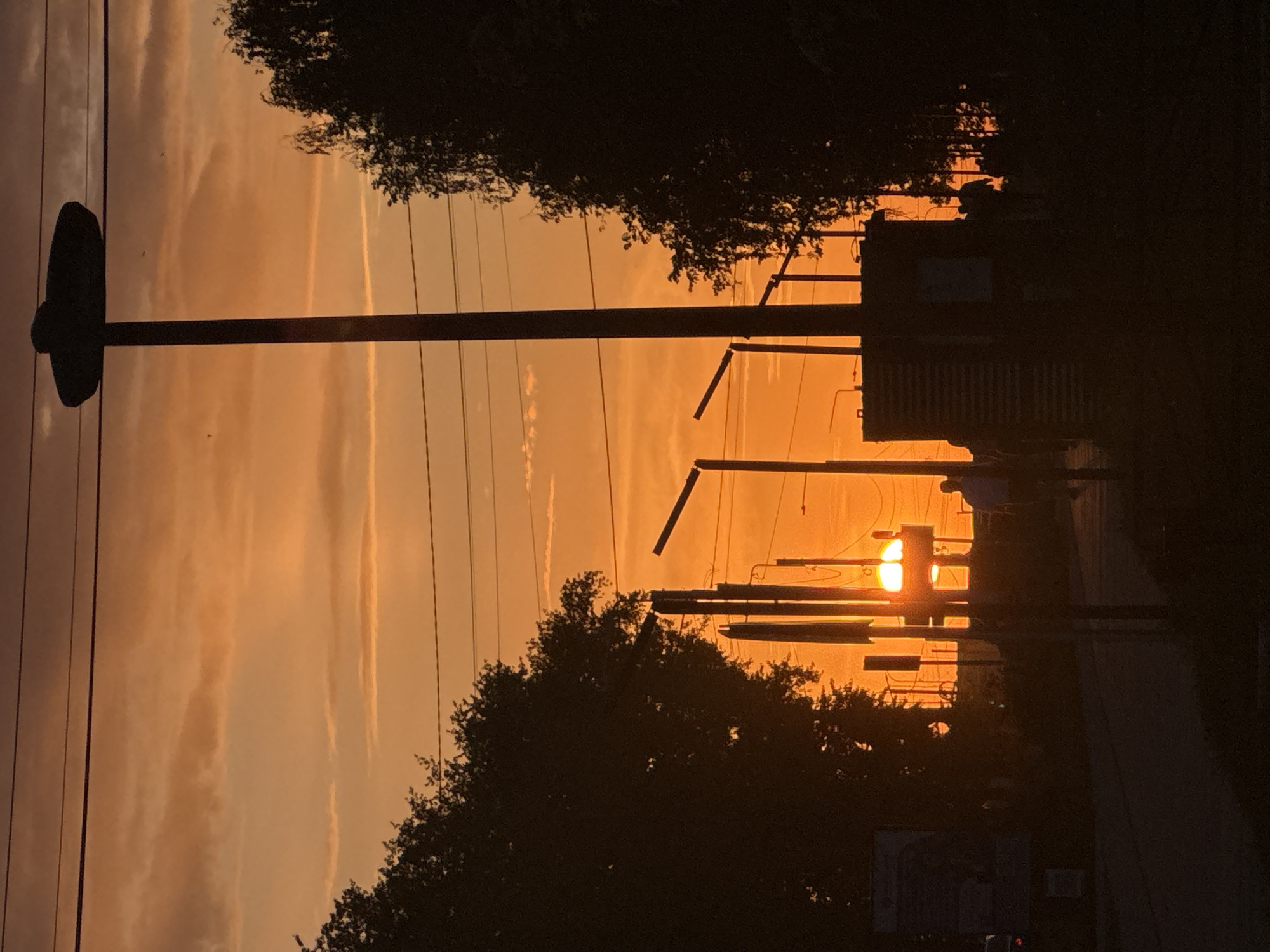 The photograph depicts a sunset in Chaikovsky, Russia. The sun is visible between the silhouettes of power lines and trees, casting an orange glow across the sky. A basketball hoop is visible on the left side of the frame. A road and some buildings are visible on the right side.
