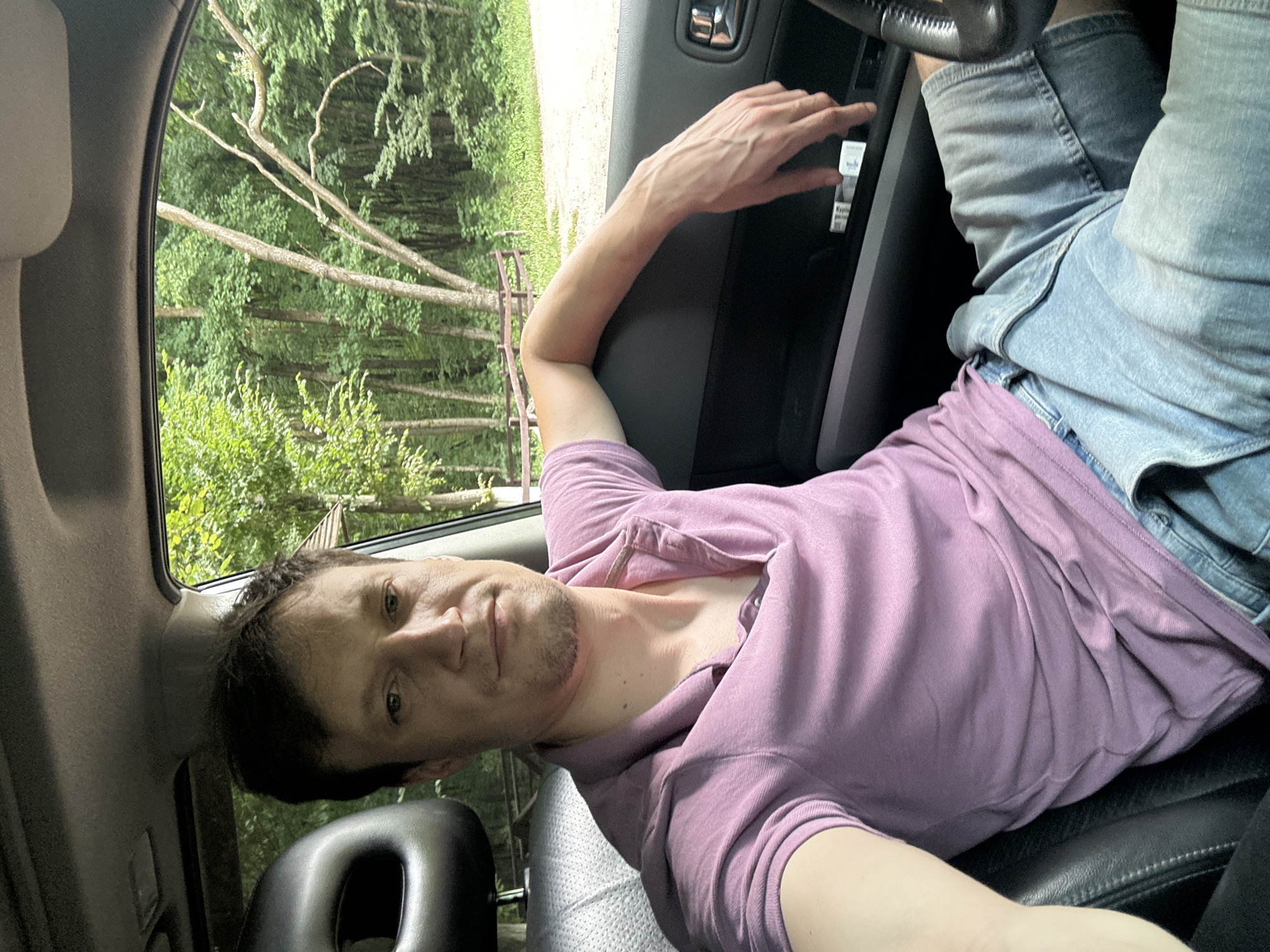 A man is sitting in the driver's seat of a car, with trees and greenery visible outside the window. He is wearing a purple polo shirt and denim shorts. His left arm is resting on the car door. The car interior shows a black headrest and part of the steering wheel. The background suggests a rural or wooded area. The image is taken from a selfie perspective.