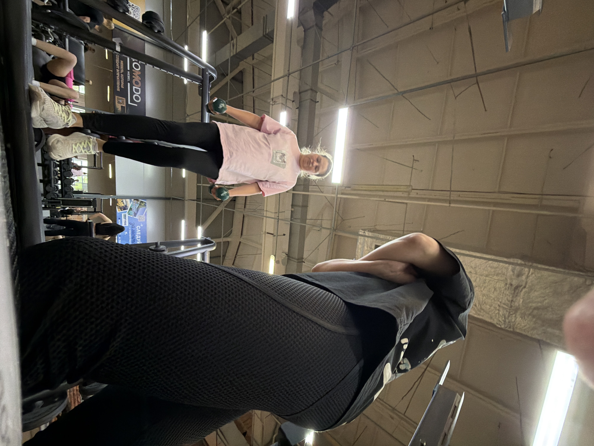 The image was taken indoors, possibly in a gym or fitness center in Chaikovsky, Russia. A person, presumably female, stands in the foreground, wearing black workout leggings, a black t-shirt, and a hoodie, with arms crossed. A second person is visible in the background, wearing a pink t-shirt and black leggings, holding green dumbbells. The background is blurry but suggests gym equipment and an industrial-looking ceiling with fluorescent lights.