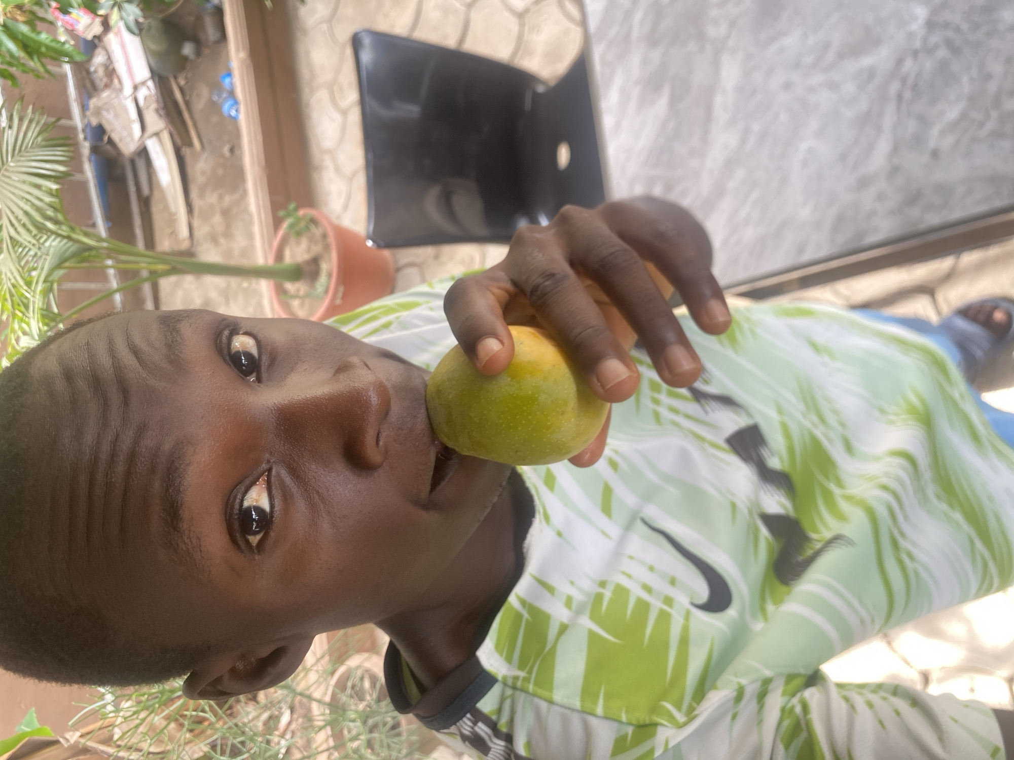 A man with dark skin and short hair is holding a small, green and yellow mango to his mouth. He is wearing a green and white patterned Nike shirt. The background includes a potted plant, a black chair, and what appears to be a paved surface. The location is Barden Yana, Nigeria.