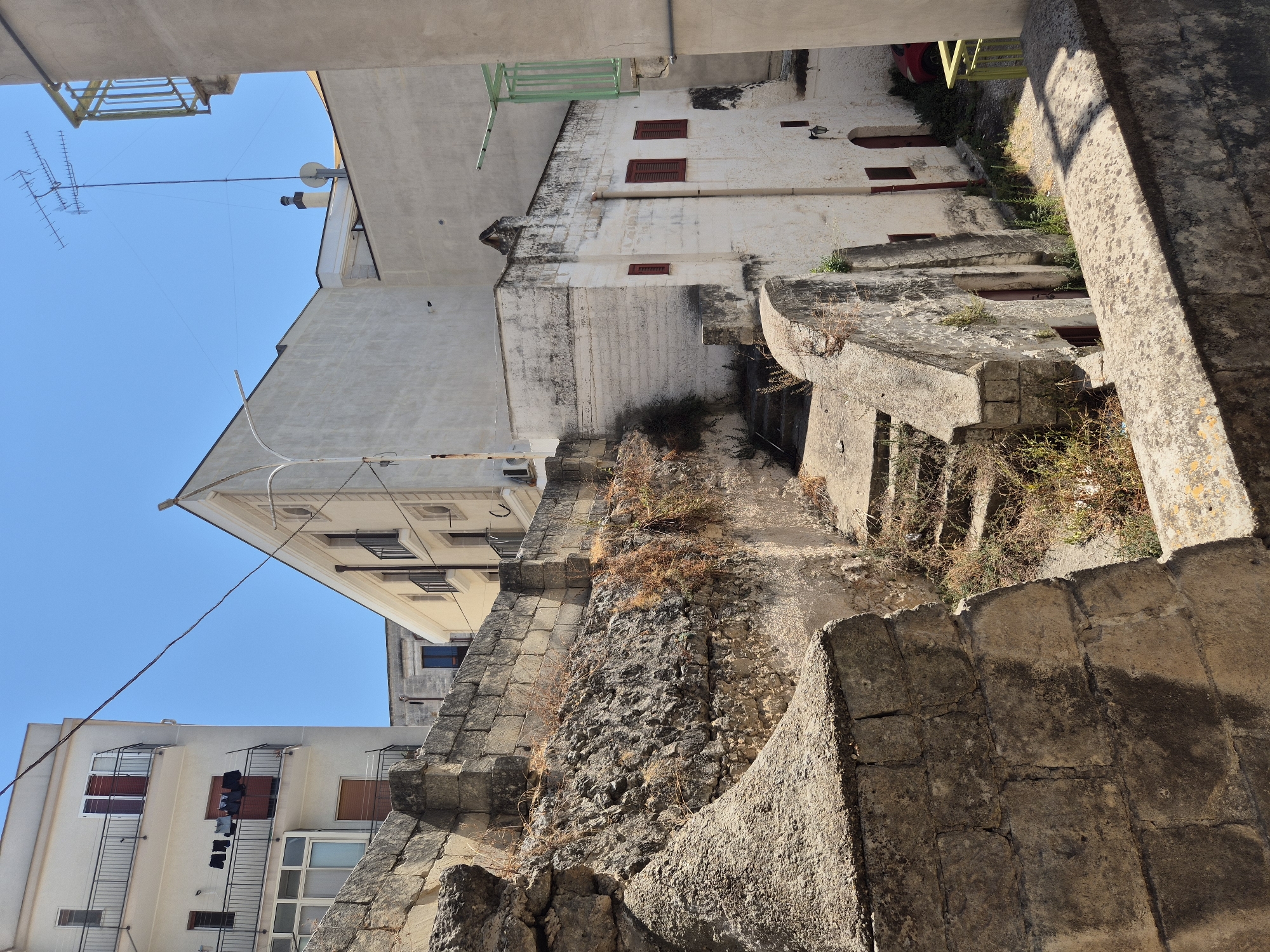 The image depicts a sun-drenched alleyway in the historic center of Laterza, Italy, characterized by its ancient stone architecture and multi-level buildings. In the foreground, a weathered stone wall forms a barrier, with the texture of rough-hewn blocks visible. Beyond this, a stone staircase descends into a lower part of the alley, bordered by more stone structures and patches of dry, overgrown vegetation.

The middle ground features a white, two-story building with red-shuttered windows and a dark archway on the lower level. A red downspout runs vertically along its side. Adjacent to this, an older stone structure with rougher textures and sparse vegetation clinging to its walls adds to the historic ambiance.

In the background, a modern, pale yellow apartment building rises on the left, with balconies featuring black railings. Laundry is hung out to dry on one of the balconies, indicating ongoing daily life. Further back, a white building with a triangular roofline and arched windows is visible. The sky above is a clear, bright blue.

The overall scene conveys a sense of timelessness and the enduring nature of the old Italian town, juxtaposing ancient stonework with more recent residential structures. The sunlight casts strong shadows, highlighting the textures and forms of the architecture.