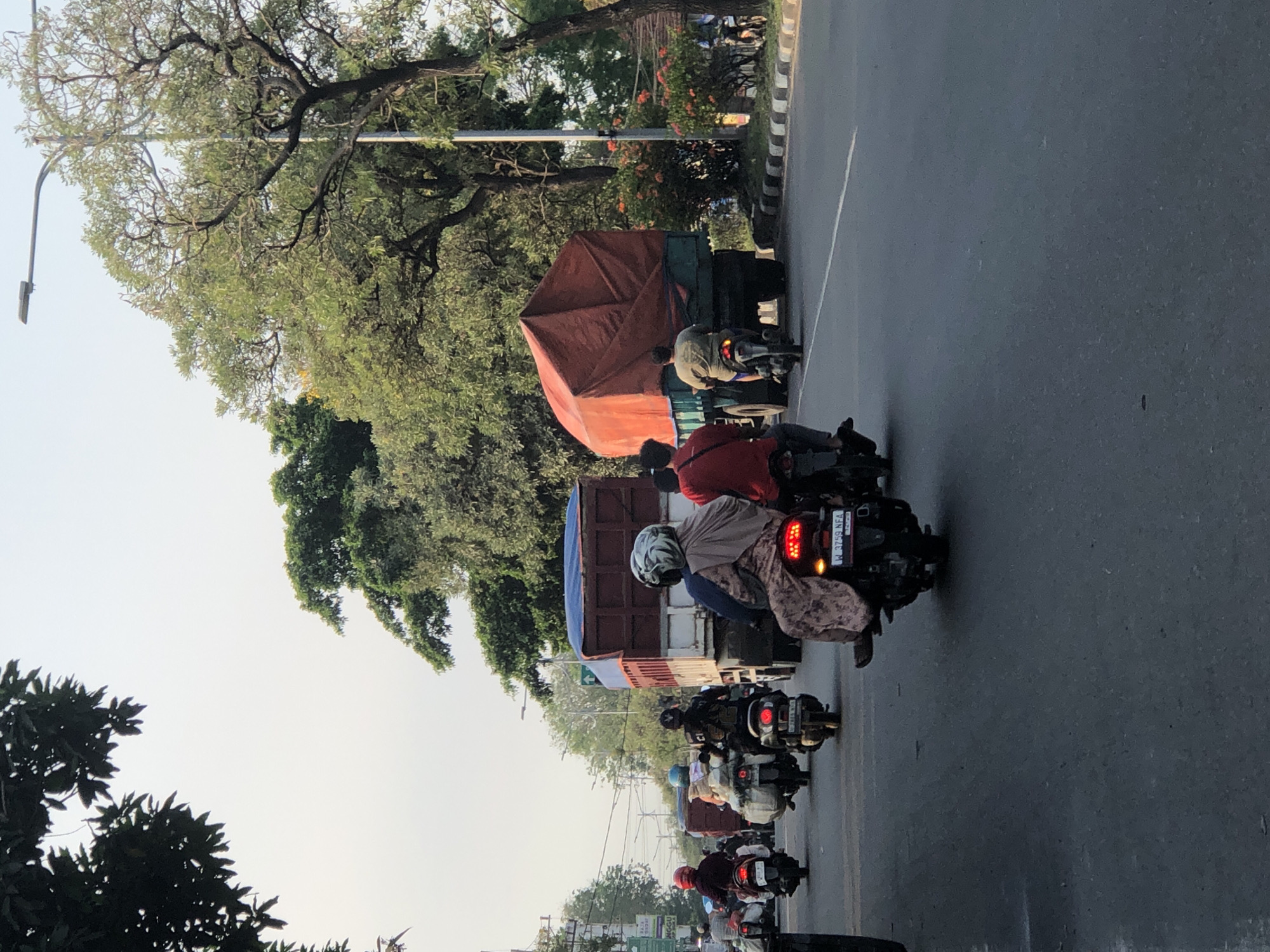 In Kludan, Indonesia, a street scene unfolds with several motorcycles and two large trucks. The motorcycles are moving in the same direction as the trucks on an asphalt road. On one motorcycle in the foreground, a person wearing a patterned brown dress and a white helmet is riding. Another motorcycle behind them has a person with a dark head covering and a patterned garment, also wearing a helmet. In front of them, two more motorcycles are visible, one with a person in a red shirt and the other with a blue head covering.
Further ahead, a large truck with a blue tarp-covered cargo is moving. To the right of this truck, another larger truck is visible, its cargo covered with a bright orange tarp. The road is lined with trees on the right side, and a street light is present. The sky is overcast with a light grey hue. The overall scene depicts a typical day in a busy Indonesian street with moderate traffic.