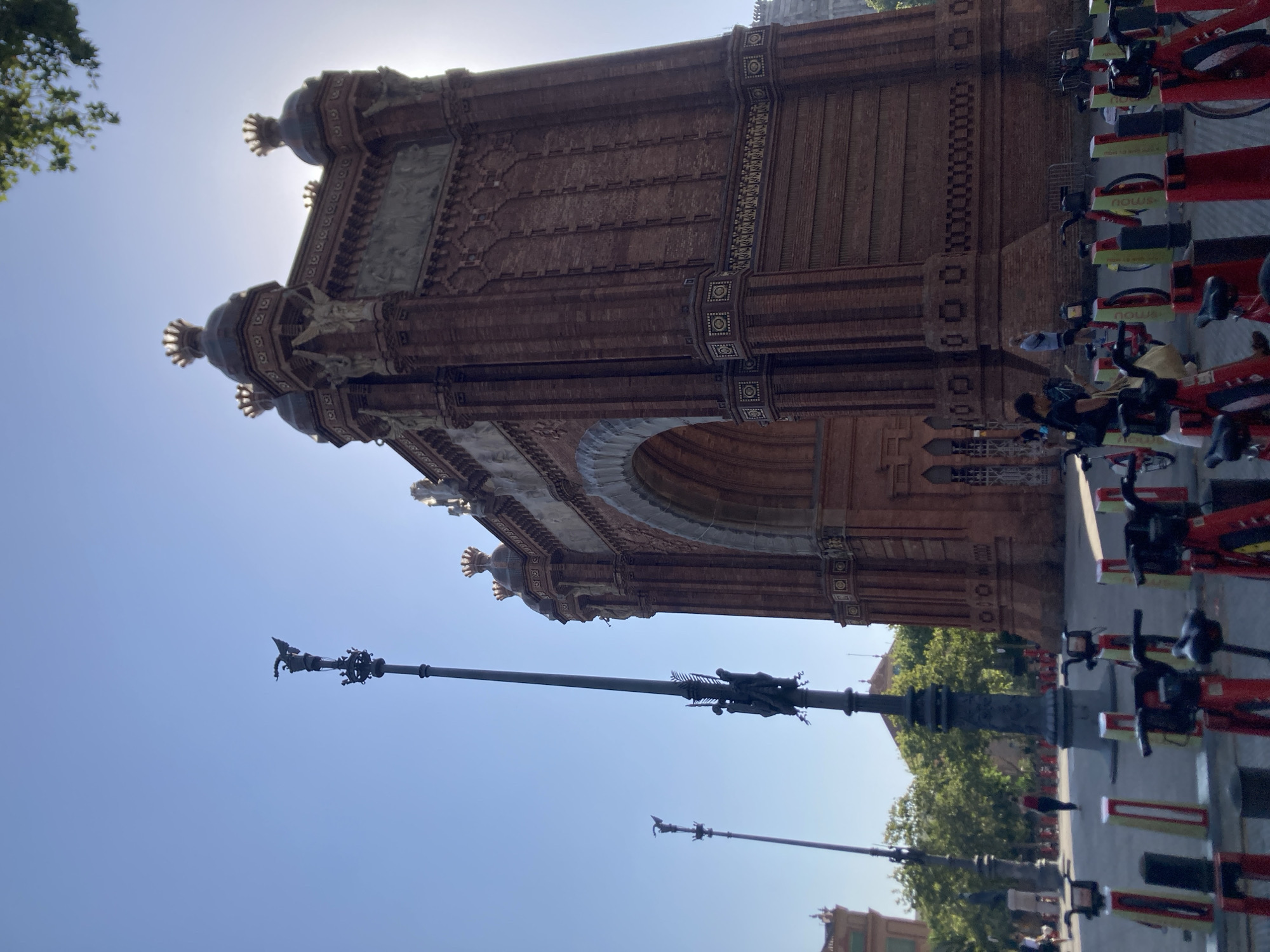 The image shows the Arc de Triomf in Barcelona, Spain, on a bright, sunny day with a clear blue sky. The monument is constructed of red brick and features intricate architectural details, including statues and decorative friezes. In the foreground, a row of red and yellow rental bicycles are parked in a docking station, suggesting a public transportation hub. A few people are visible in the background, some walking and one standing near the arc. Tall, ornate lampposts with bird-like sculptures stand to the left of the arc, one of which is closer to the camera and features more detailed figures. Trees line the background, providing a touch of greenery to the urban landscape. The sun is shining brightly, partially obscuring the top of the arc and creating a slight lens flare.