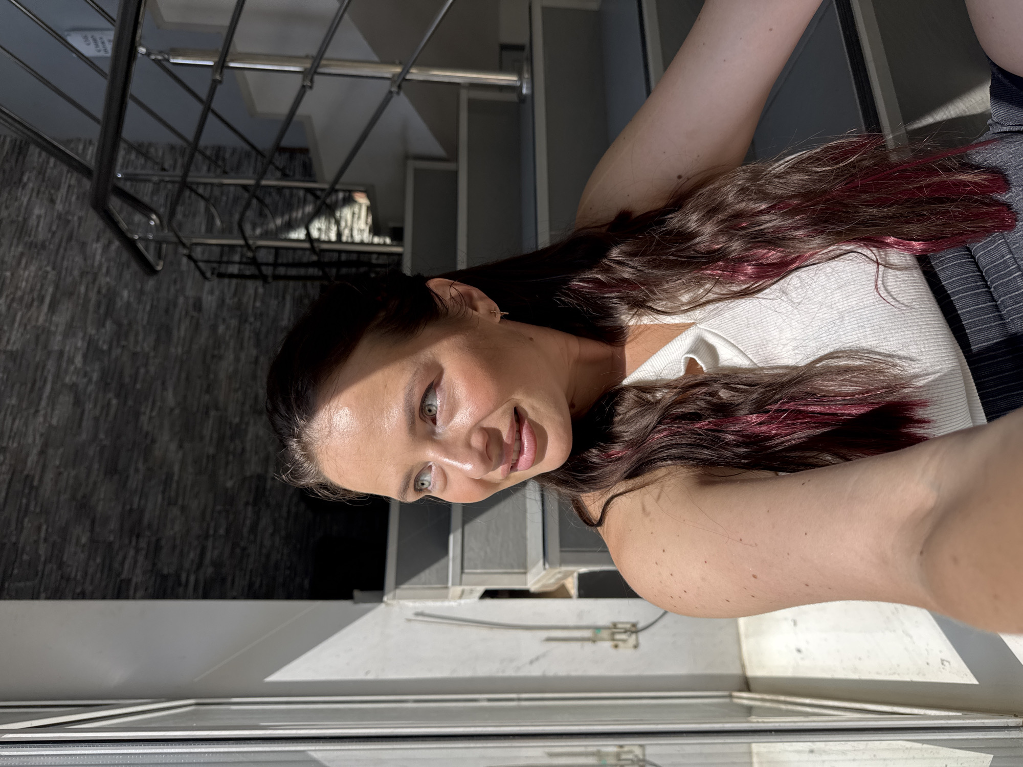 A medium shot captures a woman with long, dark hair with pink streaks sitting on a staircase. She is wearing a white tank top and a dark striped skirt. The woman is looking to her left, with sunlight illuminating her face. The staircase is made of concrete with a metal railing. The background features a textured, dark stone wall. The scene appears to be outdoors, possibly in a courtyard or an open-air building.