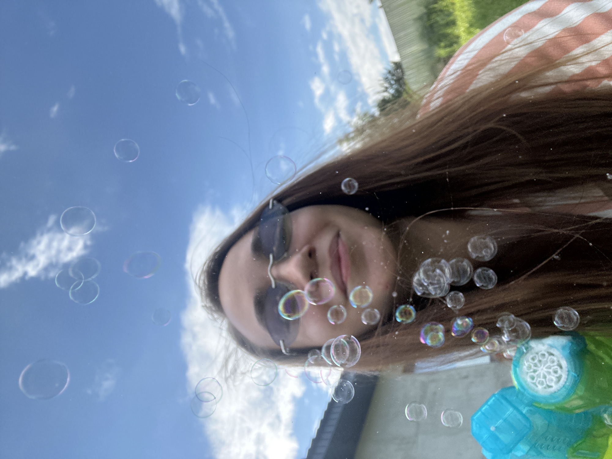 The image depicts a young woman with long brown hair, wearing sunglasses and a striped shirt, outdoors with numerous soap bubbles floating around her. She is smiling and looking upwards, towards the sky filled with clouds. In the foreground, a portion of a bubble gun can be seen. The background reveals a fence and greenery, suggesting a residential area. The scene is set in a backyard or garden, possibly in the village of Kharnavy, Russia, given the context of the request.