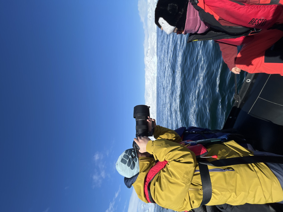The scene depicts two individuals in a black rigid inflatable boat (RIB) on dark blue, choppy water under a clear, bright blue sky with sparse white clouds. In the distance, a vast expanse of snow-capped mountains or landmass is visible. The location is a maritime environment in a polar or subpolar region, specifically in the city of Unknown, Unknown.
One individual, positioned on the left and facing right, wears a chartreuse or olive-yellow hooded jacket, a red life vest, and a light blue beanie with a dark blue graphic. This person is actively holding a professional-grade camera with a large, black telephoto lens, aiming it towards the distant landscape and water. Their face is partially obscured by the camera. The second individual, on the right, is seated and faces towards the right, away from the first person's direct line of sight. This individual wears a red life vest over red or maroon cold-weather clothing and a distinctive black and white penguin-themed hat, with a pink neck gaiter covering their lower face. Both individuals are dressed in cold-weather attire suitable for the environment. No direct interaction between the individuals is evident.