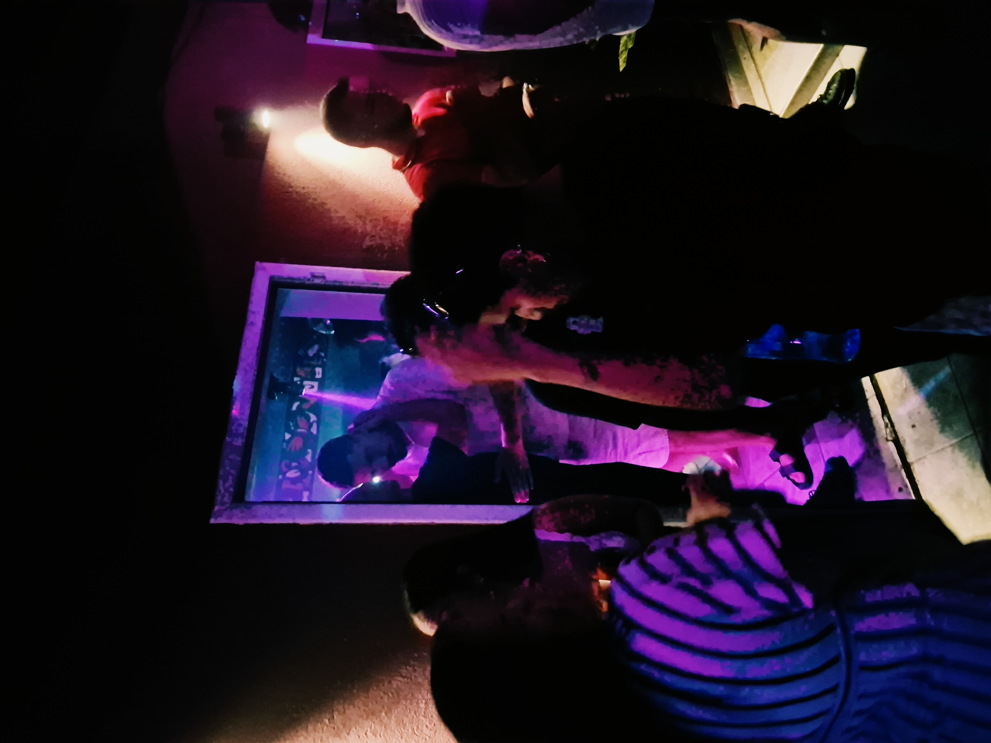 The image shows a group of people in a dimly lit indoor space with purple and pink lighting, likely a nightclub or bar. The overall ambiance is dark and somewhat hazy. In the center of the frame, a man in a white shirt and white shorts is embracing a woman. To the left, a woman in a striped top is visible from the back. Further to the right, a man in a red shirt is standing. The floor appears to be tiled. The setting is not identifiable as being in Herceg Novi, Montenegro.