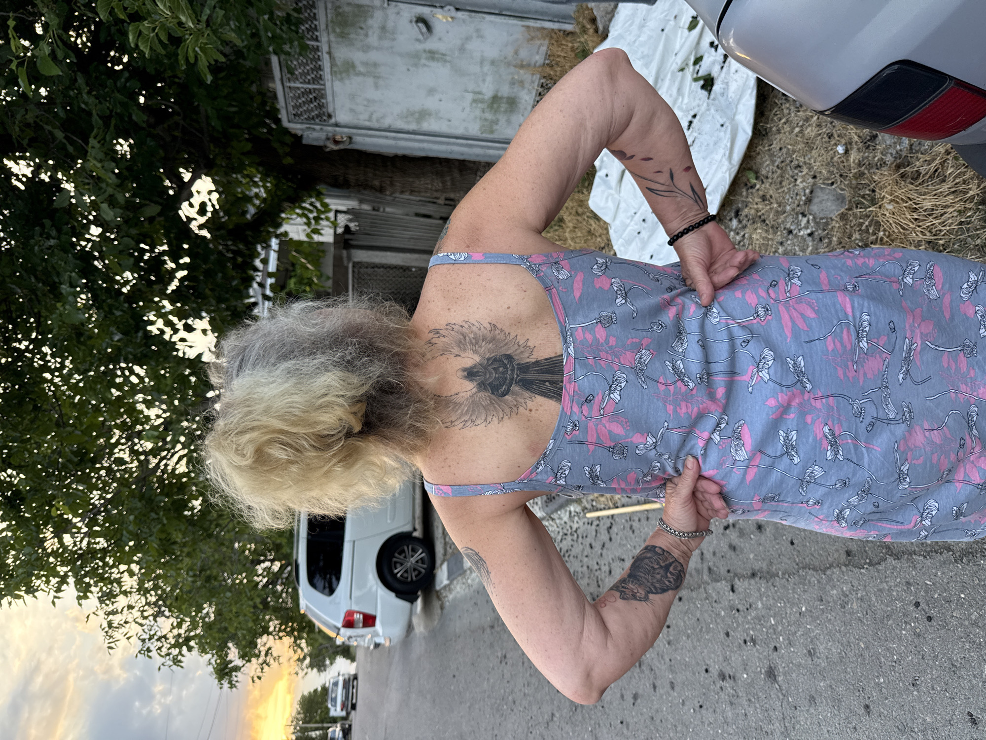 An overhead shot captures a woman from the back, standing on a paved road at dusk. Her blonde and gray hair is tied up. She is wearing a light blue spaghetti-strap dress with a pink floral pattern. She has a large tattoo of an angel on her upper back, and tattoos on both her arms. Her left hand rests on her hip, and her right hand is behind her back. In the background, a white van is parked on the side of the road. The sky is a soft orange and yellow from the setting sun. Trees and a fence are visible behind the woman. The scene appears to be in a residential area, likely in Novorossiysk, Russia.