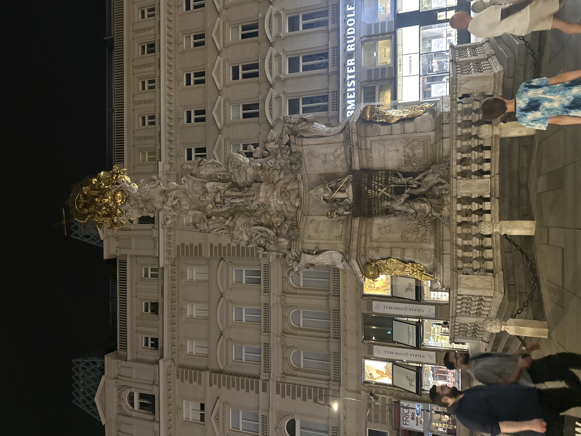 This is a nighttime, eye-level, medium shot of the Pestsäule (Plague Column) in Vienna, Austria, located on the Graben street. The monument, a tall Baroque structure adorned with sculptures and topped with a golden depiction of the Holy Trinity, is the central subject. To the left of the monument, two men are walking. The man in the foreground on the left is wearing a dark blue shirt and black pants, while the man beside him is wearing a grey short-sleeved shirt and black pants. Behind them, a building facade with multiple windows and architectural details is illuminated. Below the monument, at street level, are shops with illuminated signs, including "RMEISTER RUDOLF," "ZENITH," and "HOBNER." A young boy wearing a blue and white tie-dye t-shirt and light-colored shorts is walking away from the camera, towards the monument. To the far right, a man in a white t-shirt and light-colored shorts is walking away from the camera, and a portion of another person is visible. The ground is paved, and a chain fence surrounds the base of the monument. The sky above is dark, indicating nighttime.