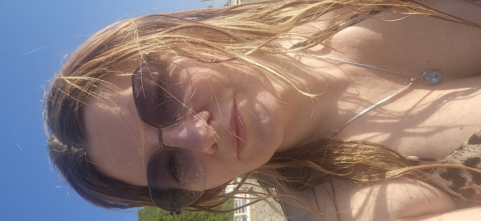 A woman is captured in a close-up selfie, taken outdoors under a clear blue sky. She is wearing sunglasses with a dark tint and a leopard-print bikini top. A silver necklace with an oval pendant rests on her collarbone. Her wet, blonde hair is windswept, with strands falling across her face and shoulders. Sunlight casts dappled shadows on her skin and hair. In the background, out of focus, are hints of greenery and a stone wall, suggesting a coastal or park-like setting. The image does not provide any specific visual cues that can identify the location as Novomikhaylovsky, Russia.