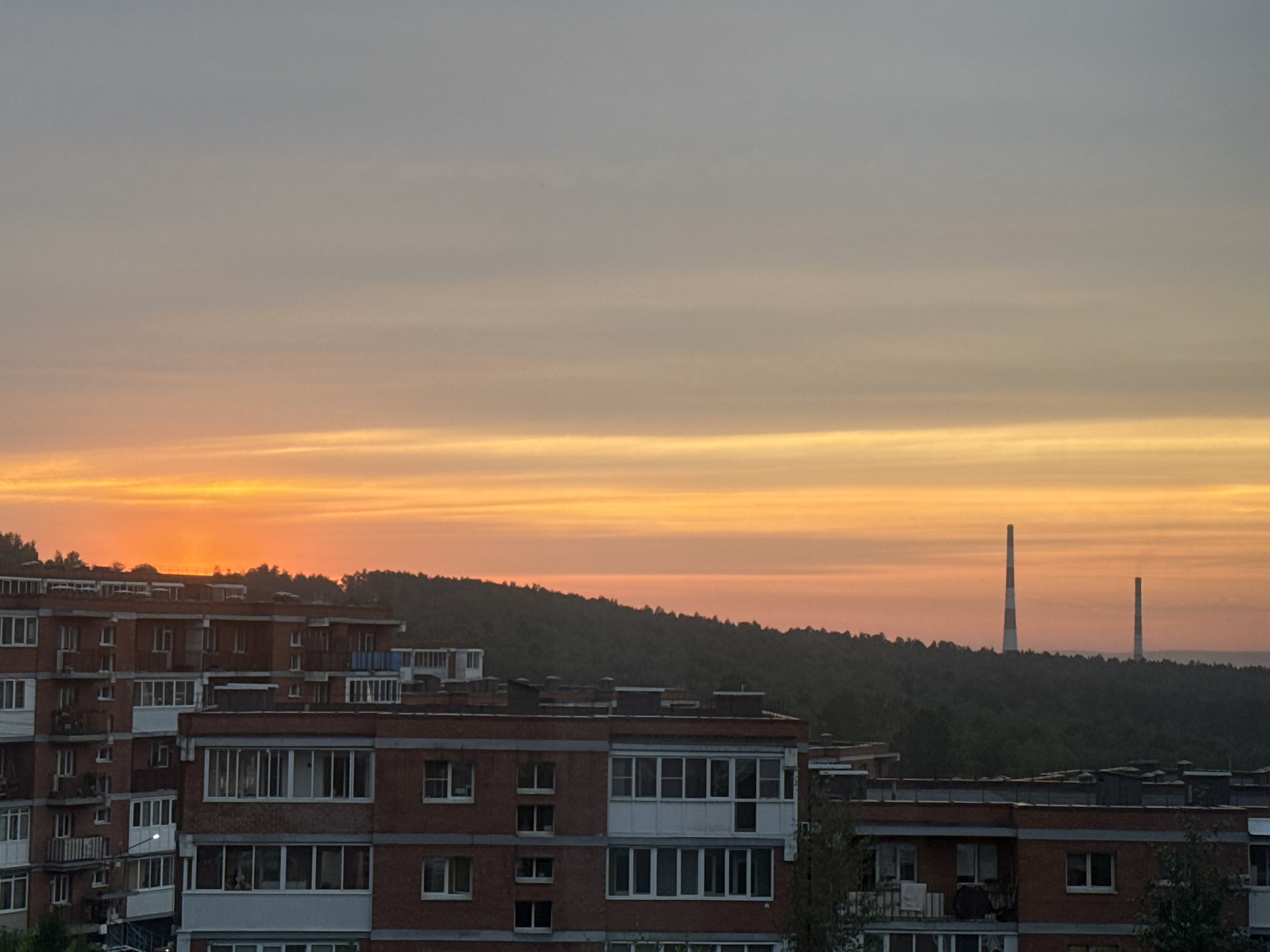 This image depicts a sunset over the city of Markova, Russia. The sky is a gradient of orange and yellow hues, with lighter shades of grey towards the top. Below the sky, a dense line of dark green trees forms a silhouette of a forest or hills. In the foreground, several multi-story brick apartment buildings are visible, characterized by their repetitive window patterns and balconies. Two tall industrial chimneys, one taller than the other and both with red and white striped markings, stand prominently above the tree line in the distance. The lighting suggests it is either dawn or dusk.