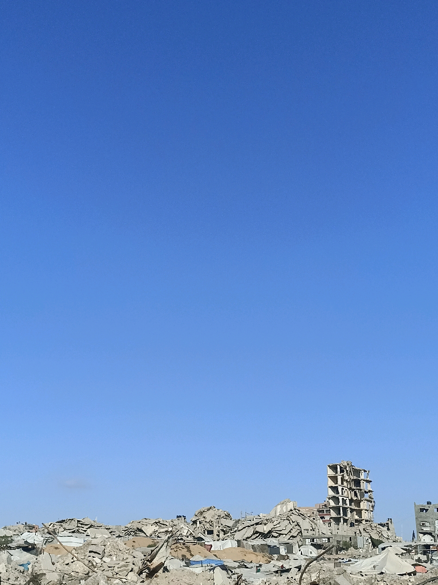 The image shows a vast expanse of rubble and debris under a clear blue sky, indicating the aftermath of significant destruction in the city of Gaza, Palestinian Territories. In the foreground, piles of concrete, twisted metal, and shattered building materials dominate the landscape, suggesting collapsed structures. Among the debris, tents and makeshift shelters are visible, hinting at the displacement of people. Further in the background, the skeletal remains of several multi-story buildings stand partially intact, their floors and walls exposed and damaged. One prominent building, a tall, concrete structure, is severely compromised, with large sections of its facade missing and its interior floors visibly broken. The scene is barren and desolate, with no visible signs of life. The environment is characterized by the widespread destruction of the urban infrastructure.