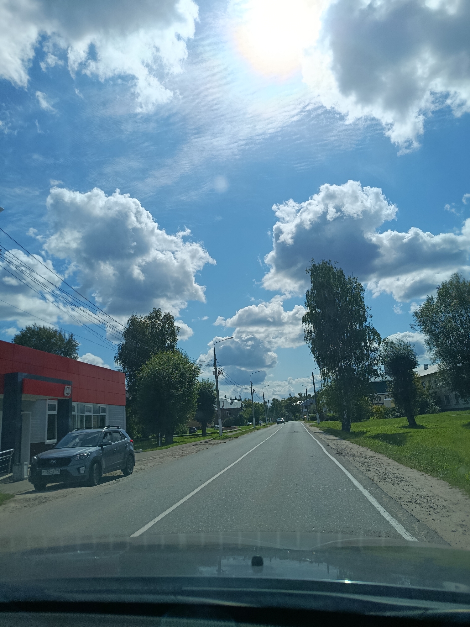 The image depicts a street scene in Verea, Russia, on a sunny day with a bright blue sky and scattered white clouds. The perspective appears to be from inside a vehicle, looking forward.
In the foreground, the dashboard of a car is visible. To the left of the road, there is a modern-looking building with a red facade and a grey lower section, featuring large windows. A dark grey SUV is parked in front of this building, facing the street.
The road itself is paved and has white lane markings. Trees line the sides of the road, with some appearing taller and fuller than others. In the distance, a few houses and other buildings can be seen, suggesting a residential or semi-rural area. A few cars are visible driving on the road in the distance. Electrical wires are strung overhead, supported by poles. The overall impression is a calm, everyday scene in a Russian town.