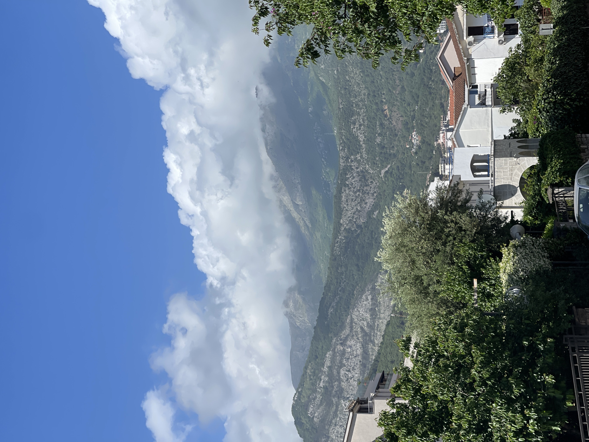 The image, taken at a slight upward angle, shows a view in Budva, Montenegro, featuring a combination of natural and urban elements. The left side of the image is dominated by a bright blue sky with large, white cumulus clouds. Adjacent to the sky, a large, verdant mountain range rises, partially obscured by a light mist. On the right side of the image, several white buildings are visible, including a building with a light red roof. The buildings are interspersed with various trees and bushes, partially obscuring the view of the buildings. At the very edge of the right side of the image, a fence and a part of a car are visible.