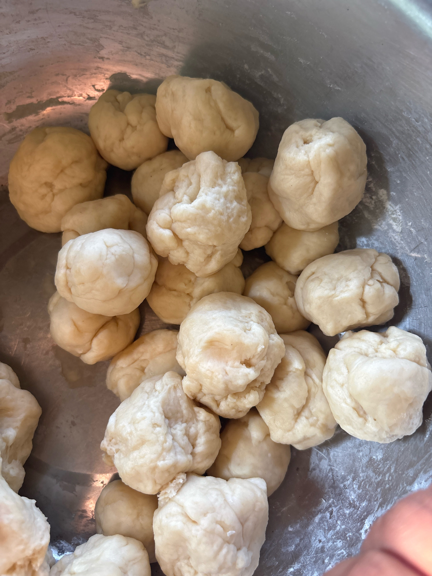 The image shows a close-up, overhead view of a metal bowl filled with roughly formed balls of dough. The dough is a pale, creamy yellow color and appears to be soft and slightly lumpy. Some of the dough balls are larger and more rounded, while others are smaller and more irregular in shape. The bowl itself is metallic and appears to be stainless steel, with some smudges and scratches visible on its surface. The lighting suggests an indoor environment, likely a kitchen, with reflections visible on the polished metal of the bowl. There are no identifiable landmarks or contextual elements within the image to indicate a specific location in Arateng, Indonesia.