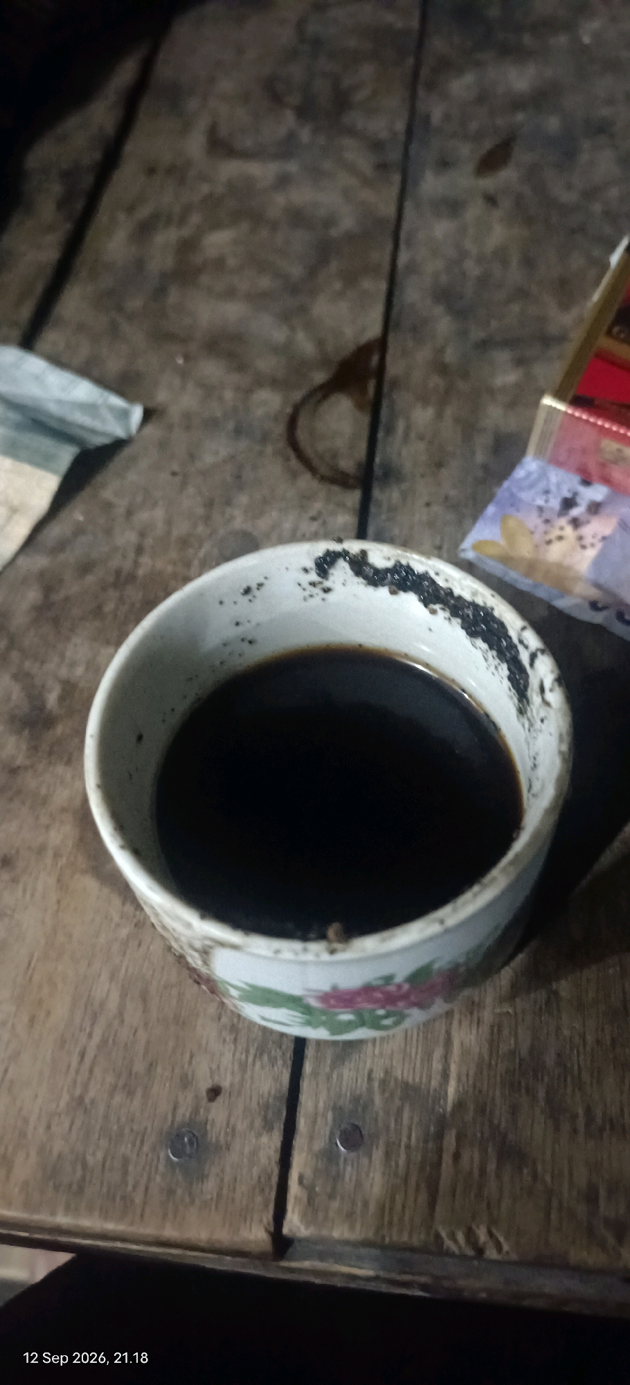 The image shows a close-up of a white ceramic cup filled with dark liquid, likely coffee, resting on a wooden surface. The rim of the cup has dried residue and some small particles. A small portion of what appears to be a pack of cigarettes or similar packaging is visible to the right of the cup. To the left, a piece of light-colored fabric or paper is partially visible. The wooden surface appears to be made of planks with visible grain and some wear. The overall lighting suggests an indoor or dimly lit outdoor setting, possibly at night, given the timestamp at the bottom left corner which reads "12 Sep 2026, 21.18". There are no people visible in the frame. The scene is set on a wooden table. The location is identified as Mojopetung, Indonesia.