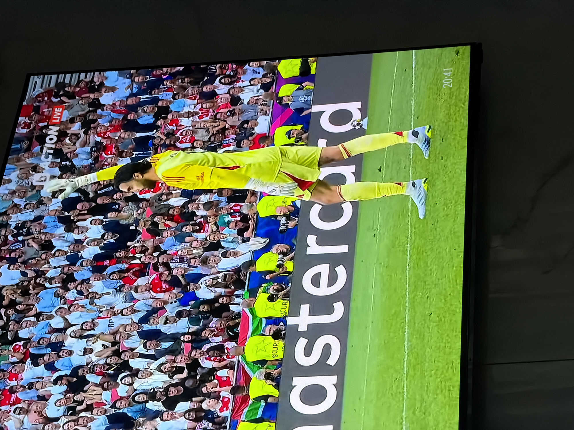 The image shows a television screen displaying a soccer game. The game appears to be a live broadcast, as indicated by the "ACTION LIVE" text in the upper left corner. The time displayed on the screen is 20:41. A soccer player in yellow goalie gear is positioned on the field. A large crowd is visible in the background. A banner with the "Mastercard" logo runs vertically along the side of the field. The image was taken inside a room, based on the wall visible in the background. There is no indication that this scene is from Asenovgrad, Bulgaria.