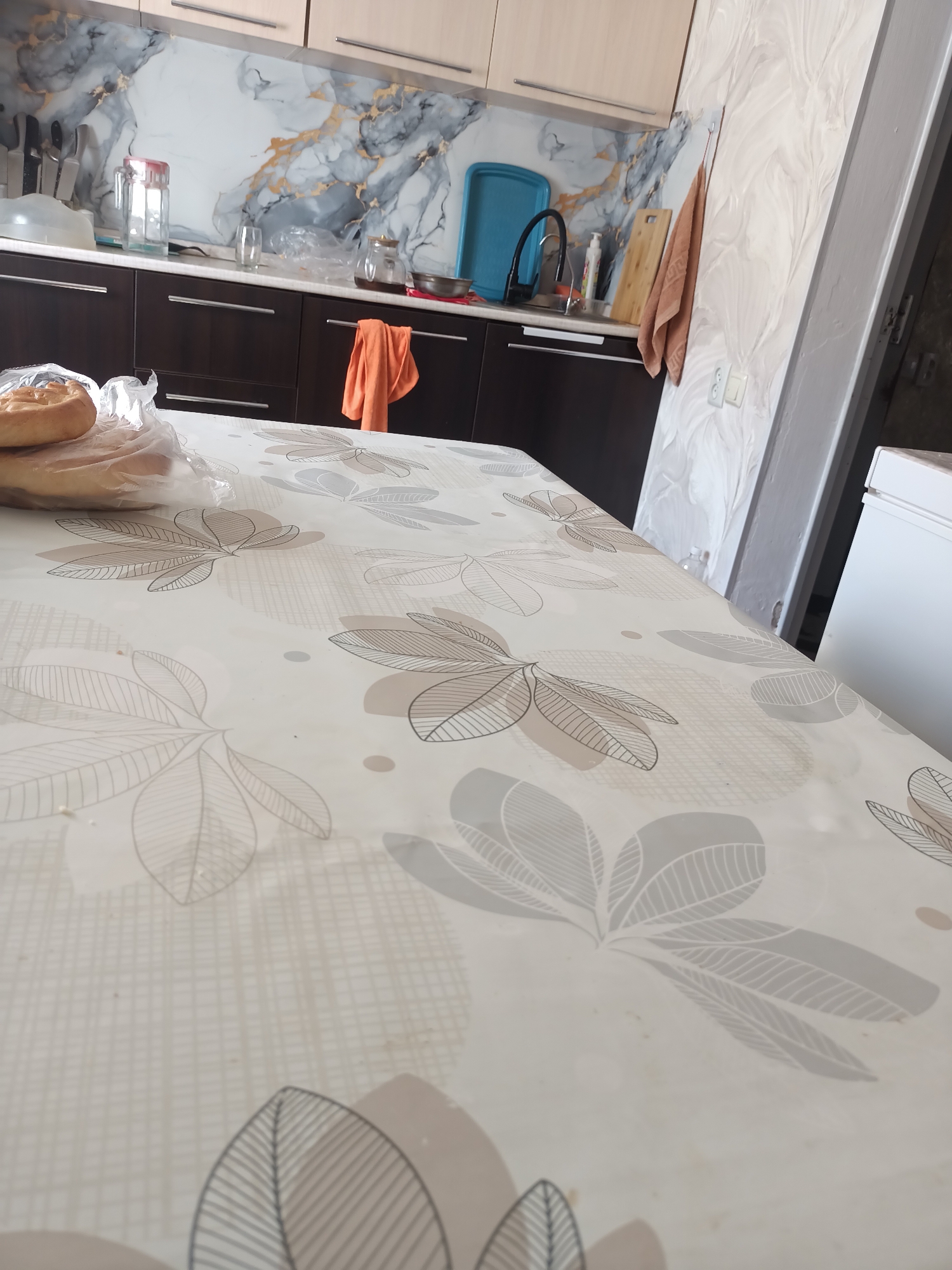 The image depicts a kitchen interior, likely in Azat, Kazakhstan. The scene is viewed from a perspective close to a table covered with a patterned tablecloth featuring leaves and circles. In the foreground, there is a loaf of bread in a plastic bag on the table.
The background reveals a kitchen counter with dark brown cabinets below. Above the counter, there's a backsplash with a marble-like pattern featuring grey, white, and gold veins. Various kitchen items are visible on the counter, including a knife block with knives, a glass, a pitcher with a red lid, a smaller pitcher with a dark liquid, a blue tray, a sink with a faucet, a bottle of soap, and a wooden cutting board. An orange towel hangs from a hook on the wall to the right of the sink. The wall to the far right of the image has a textured, light-colored finish. A white appliance, possibly a refrigerator, is partially visible on the right edge of the frame. The overall impression is of an occupied and functional kitchen space.