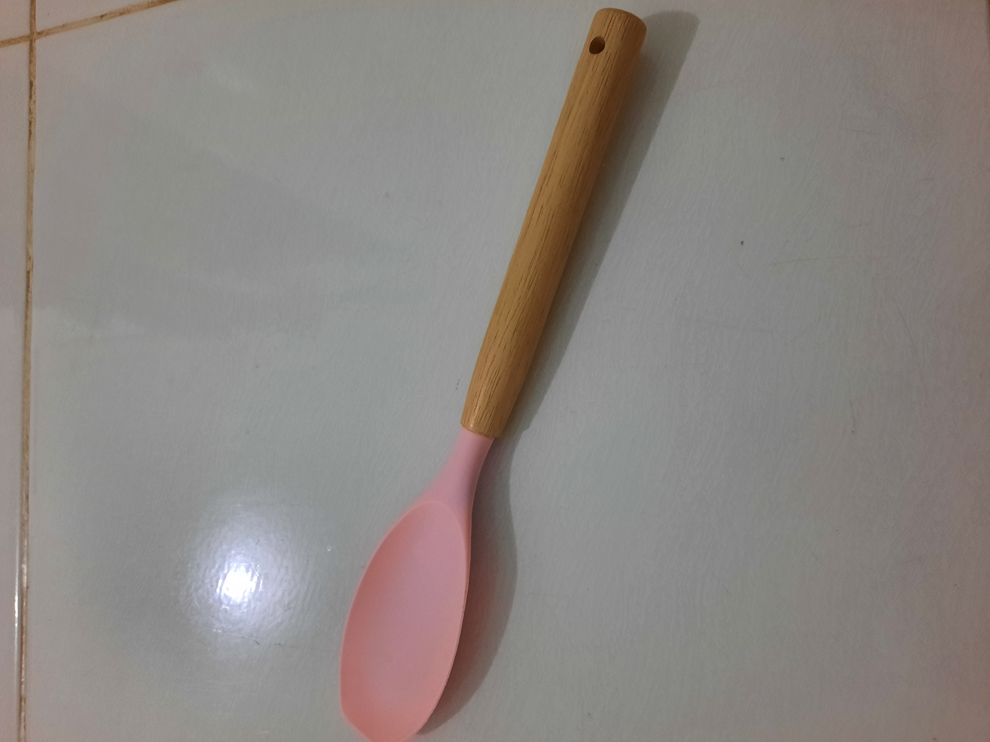 A light pink silicone spoon with a wooden handle is lying on a light gray tiled surface. The spoon is positioned diagonally, with the spoon's head on the left and the handle extending to the right. The wooden handle has a small hole at its end. The tiled surface appears to be a floor. The image was taken in Lhokseumawe, Indonesia.