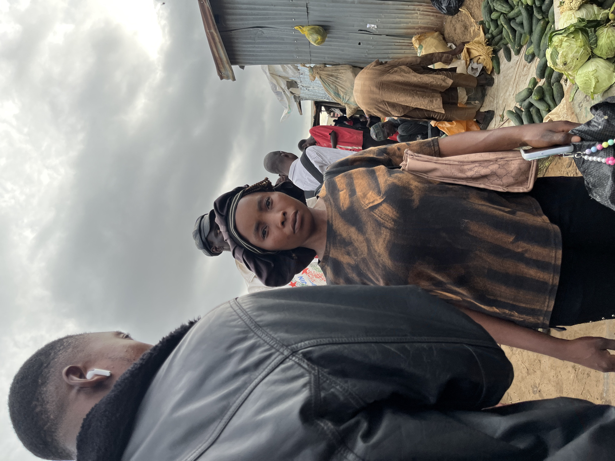 This media file captures a busy market scene, likely in Gura Top, Nigeria. The image is oriented vertically, with the sky taking up a significant portion of the upper left. The sky is overcast, with thick, grey clouds suggesting a possibly dull or rainy day. A corrugated metal roof structure is visible along the top edge of the frame, indicating a market stall or shelter.
The foreground is dominated by the shoulders and head of a person wearing a black leather jacket. To the right of this person is another individual, a woman, who is looking towards the camera. She is wearing a patterned top in shades of brown and black and has a headscarf. Her expression is neutral, perhaps slightly curious or guarded. She is holding a light brown purse with a textured pattern and a mobile phone, with a beaded string attached to it.
Behind the woman, the market unfolds. Several people are visible, some in casual clothing like a red t-shirt and a white and blue striped shirt. They appear to be standing or moving, suggesting a bustling environment. In the background, piles of produce are evident, most notably a large quantity of cucumbers and cabbages, indicating a food market. There are also sacks and bags, typical of market goods. The ground appears to be dirt or a rough, natural surface. The overall impression is one of a lively, everyday market with people engaged in commerce and daily activities.