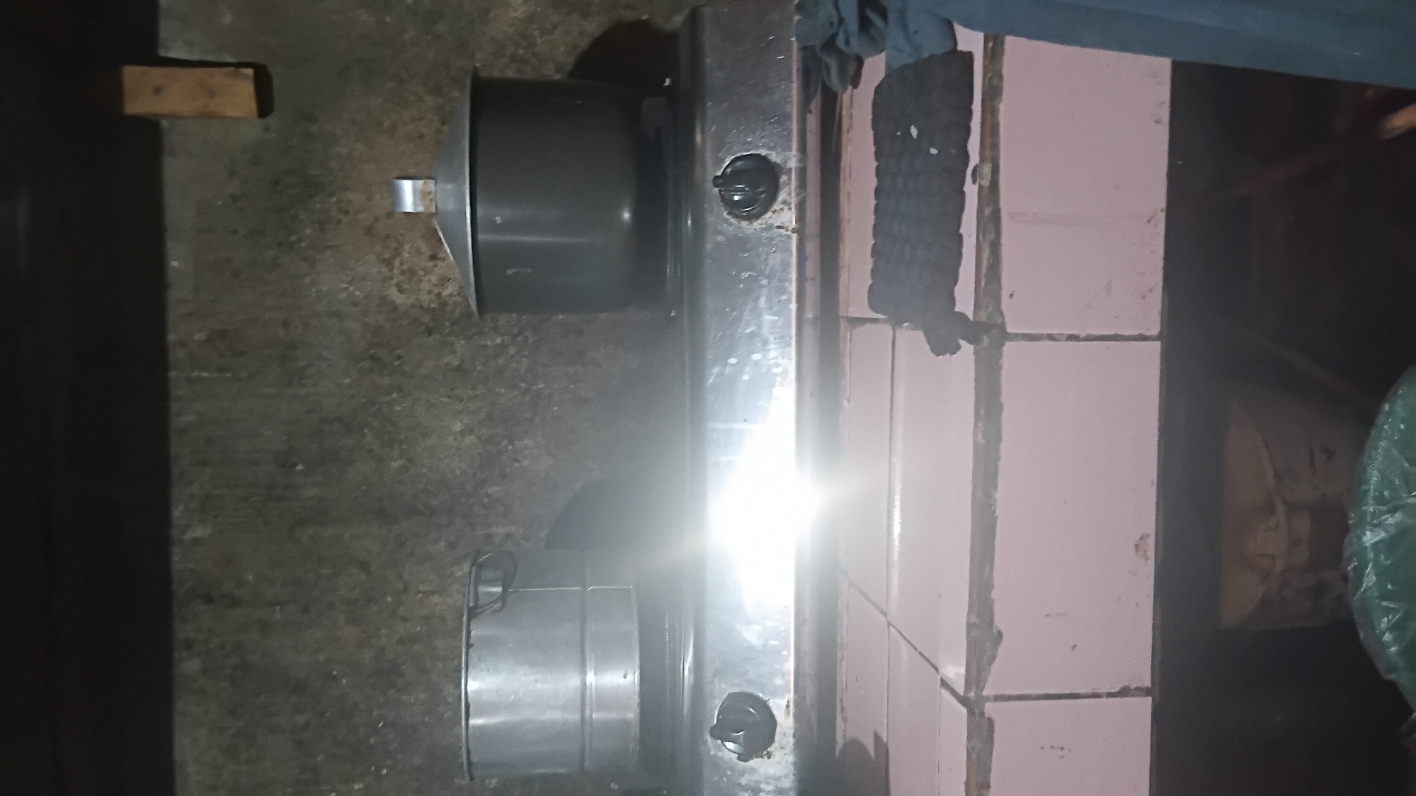 The image depicts a domestic kitchen scene in Prabumulih, Indonesia. A stainless steel gas stove with two burners is positioned on a pink tiled countertop. On the left burner, there is a silver pot, possibly a steamer. On the right burner, a dark gray lidded pot is visible. The background wall appears to be rough and gray, with a small wooden plank affixed to it on the upper right side. Below the countertop, obscured by shadows, is a glimpse of a trash can and a green plastic bag. A black woven potholder is placed on the countertop to the right of the stove, and a dark blue cloth is draped on the far right. The lighting suggests it is nighttime or a poorly lit interior.