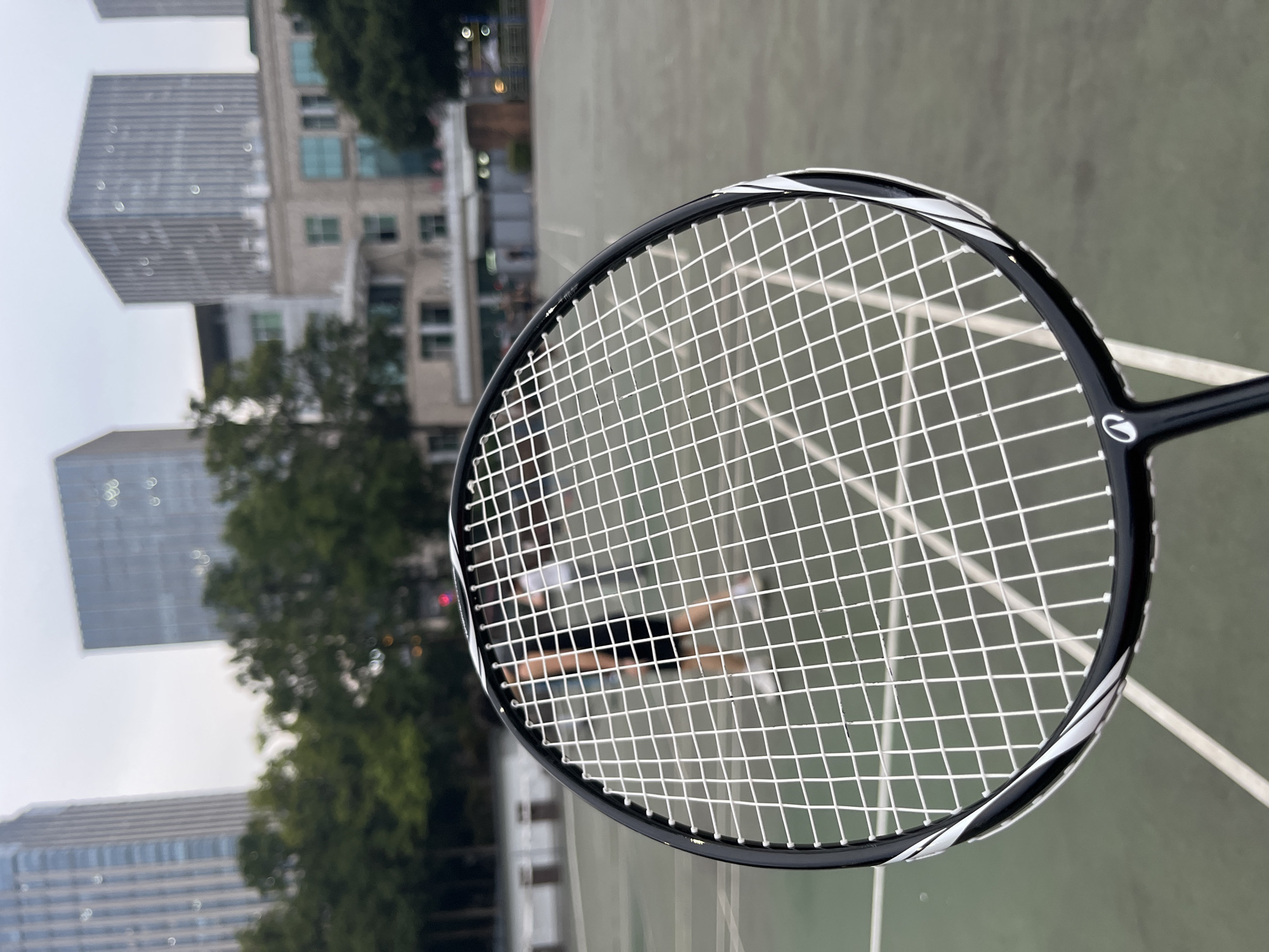 A close-up shot captures a badminton racket with its strings filling the frame, positioned over a blurred outdoor court. In the background, several tall buildings rise, some with reflective surfaces. Trees with green foliage are visible between the buildings, and hints of lights from distant structures suggest it might be late afternoon or early evening. The court appears to be made of asphalt, with white lines marking its boundaries. Through the racket's strings, a person can be seen in the middle distance, blurred and indistinct, wearing dark clothing and appearing to be playing or standing on the court. The overall scene is a blend of urban landscape and recreational activity, with the focus on the detail of the badminton racket. No visible text is present in the image.
