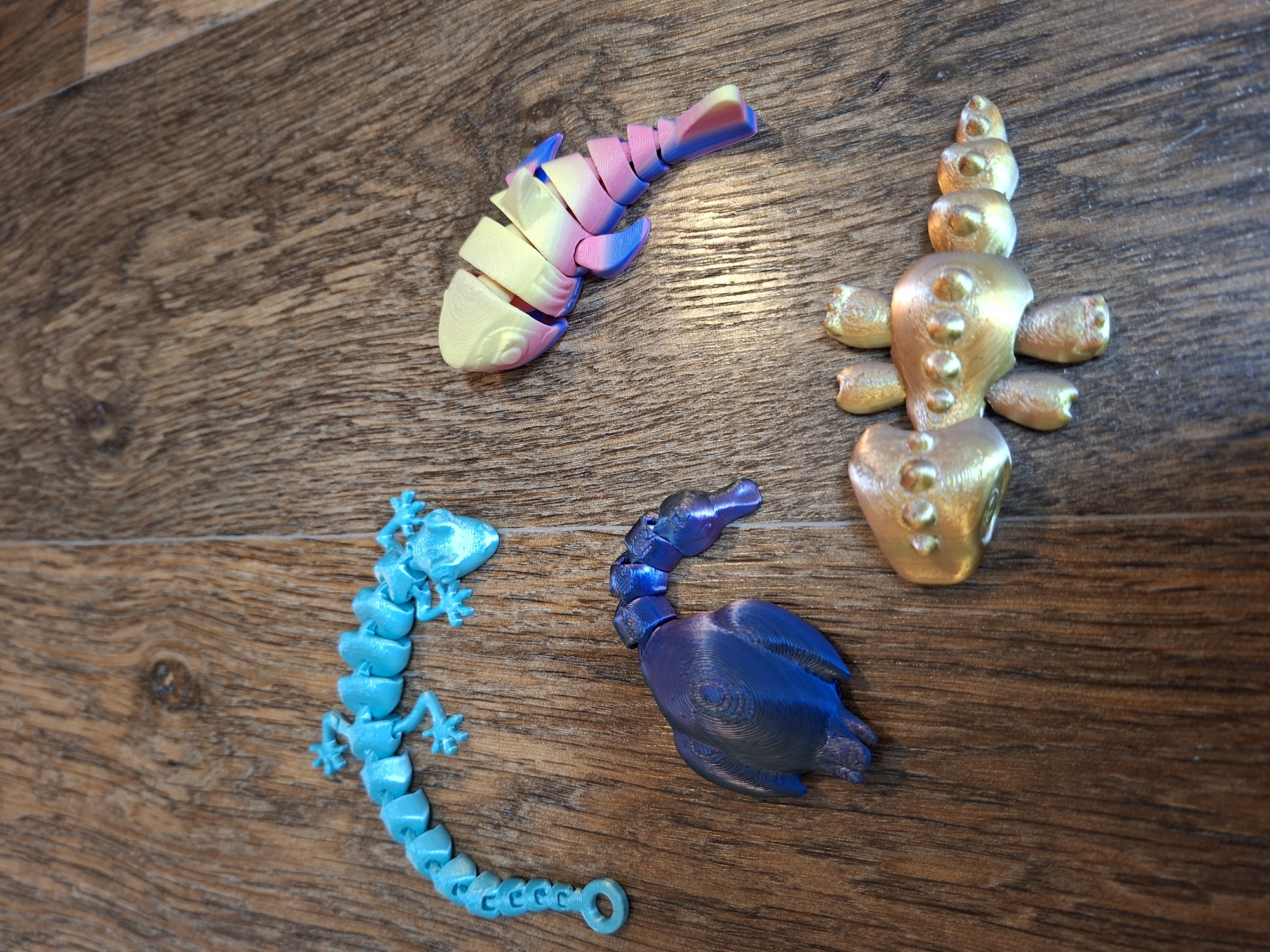 The image shows four 3D-printed articulated toys on a wooden surface. From left to right, the toys are a turquoise lizard, a multi-colored fish, a dark blue and purple bird, and a metallic gold scorpion. The wooden surface appears to be a table or floor with a wood grain pattern. There are no people or signs of human activity in the image. The image does not provide any information about the location in the city of Gorkovskiy, Russia.