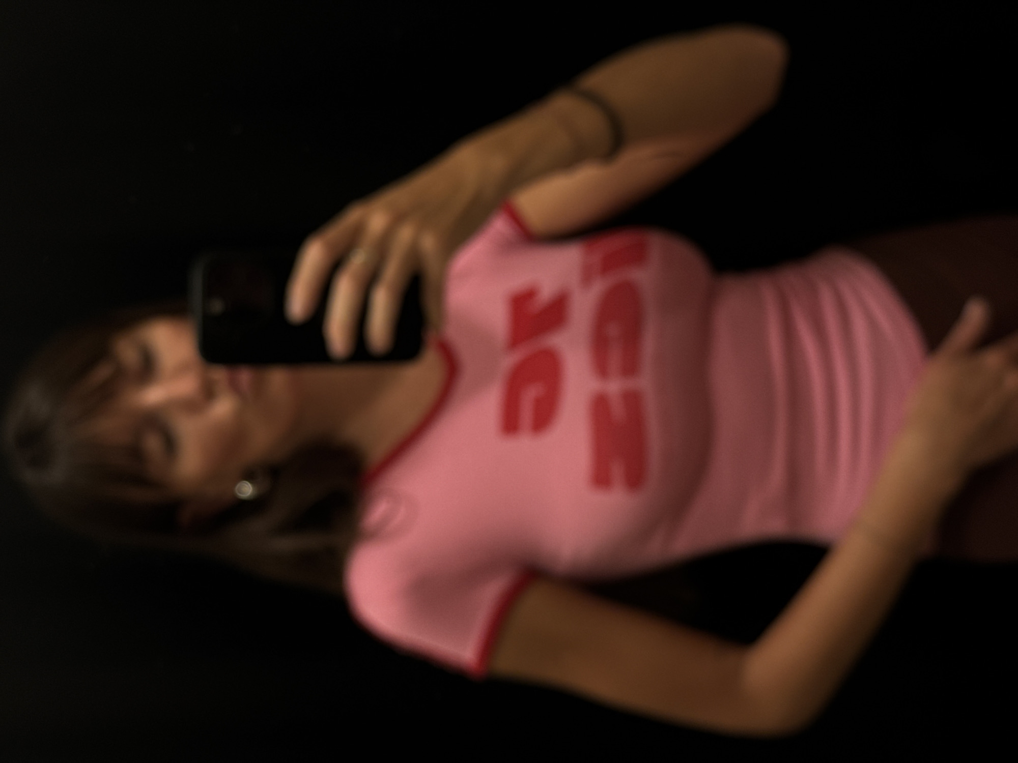 A woman is taking a selfie in a dark environment, possibly indoors. She is wearing a light pink, form-fitting top with red trim and lettering on the front that appears to be mirrored. She is holding a black smartphone up to her face. The image has poor focus. The location in Kuta, Indonesia, cannot be determined from the image.
