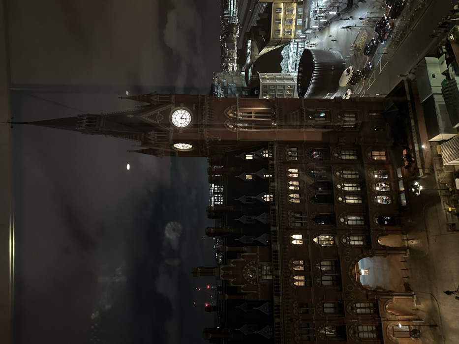 A night view showcases St. Pancras Renaissance Hotel, its clock tower illuminated under the moonlight. Its architecture is reflected on the window in the left side. To the right, a street scene reveals illuminated buildings, streetlights, and parked vehicles. The sky is dark with clouds.