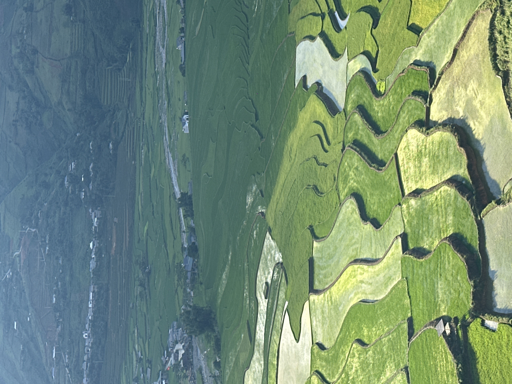 This image presents an expansive, elevated view of terraced rice fields and a valley landscape in Tú Lệ, Vietnam. The foreground is dominated by meticulously shaped, vibrant green rice terraces, some flooded with water reflecting the sky, while others are densely planted with young rice stalks. These terraces follow undulating, organic contours, separated by distinct dark earth retaining walls. A small, open-sided wooden shelter with a dark, possibly thatched roof is visible on a lower-left terrace. The midground extends into a wider valley floor, also cultivated with rice, and features a light-colored, winding river or stream bed. Scattered throughout this area are several clusters of residential structures, indicative of small villages or hamlets, with houses varying in color from white to darker tones, connected by visible roads. In the background, steep, heavily vegetated mountains rise, displaying some terracing and additional scattered buildings on their lower slopes, appearing distant and slightly hazy. No individuals or specific actions are discernible.