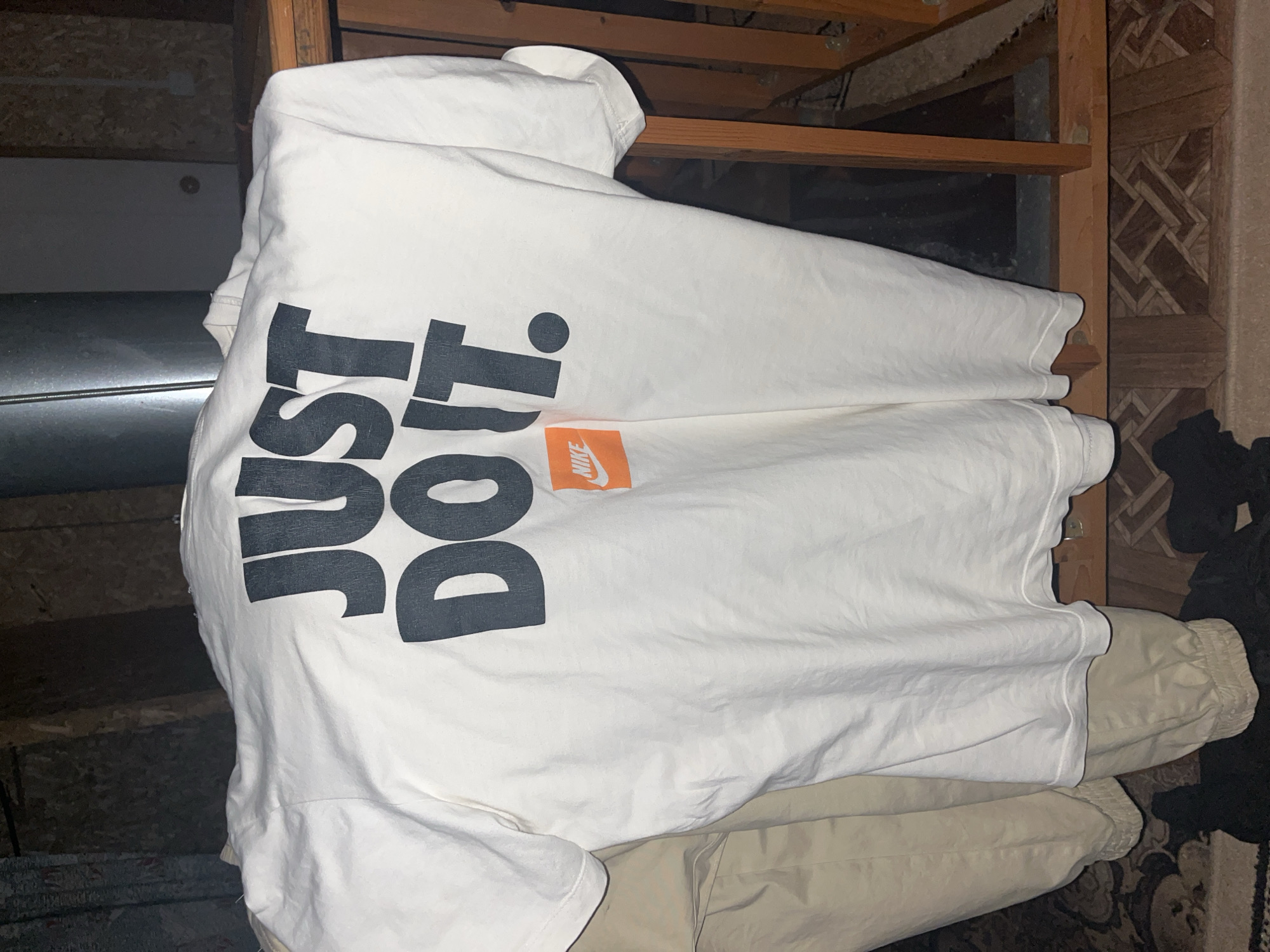 A white Nike t-shirt with the words "JUST DO IT" printed in large black letters across the back is displayed against a dark background. A small orange Nike logo is also visible on the shirt. The t-shirt appears to be hanging or draped over something, possibly in a storage area or attic, given the exposed wooden beams and insulation visible in the background. The lighting is dim, suggesting an indoor environment with limited natural light. The image is taken from a slightly elevated angle, focusing on the t-shirt. There is no discernible city or specific location information in the image itself, and therefore no connection to Chaikovsky, Russia can be made.