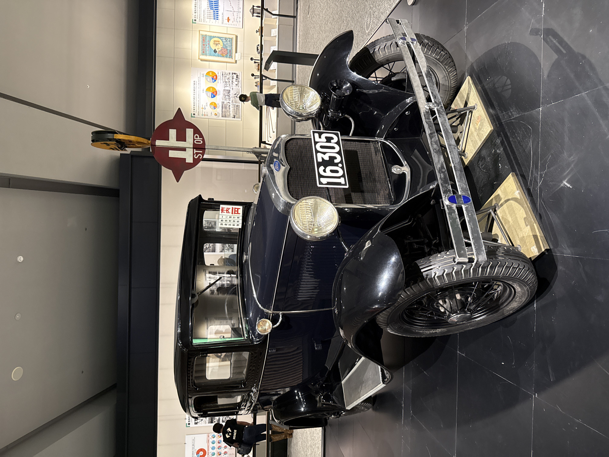 An antique dark blue car, possibly a Ford, is the central subject of this indoor photograph, displayed within what appears to be a museum or exhibition hall. The car is positioned at an angle, with its front facing towards the right side of the frame. It features classic design elements such as prominent circular headlights, a chrome grille with the "Ford" logo, and a black license plate displaying the numbers "16.305" in white. The vehicle has spoked wheels with black tires.
To the left of the car, a red and white circular stop sign is visible, featuring both the Japanese character "止" (stop) and the English text "ST OP". The setting includes a dark, polished floor, which reflects parts of the car and the room. The background consists of light-colored walls adorned with various informational posters and charts, some featuring pie graphs and diagrams. Partial OCR text from these posters indicates phrases like "■工場の出の動向" (trends in factory output) and "■ガス・電気の" (gas/electricity).
Several people are faintly visible in the background, observing the exhibit. One person wears a green shirt and a cap behind the car, while another person wearing a dark shirt with the word "PEACE" printed on the back is seen closer to the rear of the vehicle. The lighting suggests an artificial, indoor environment, emphasizing the static display of the historical automobile. No specific activity beyond viewing the exhibit is discernible.