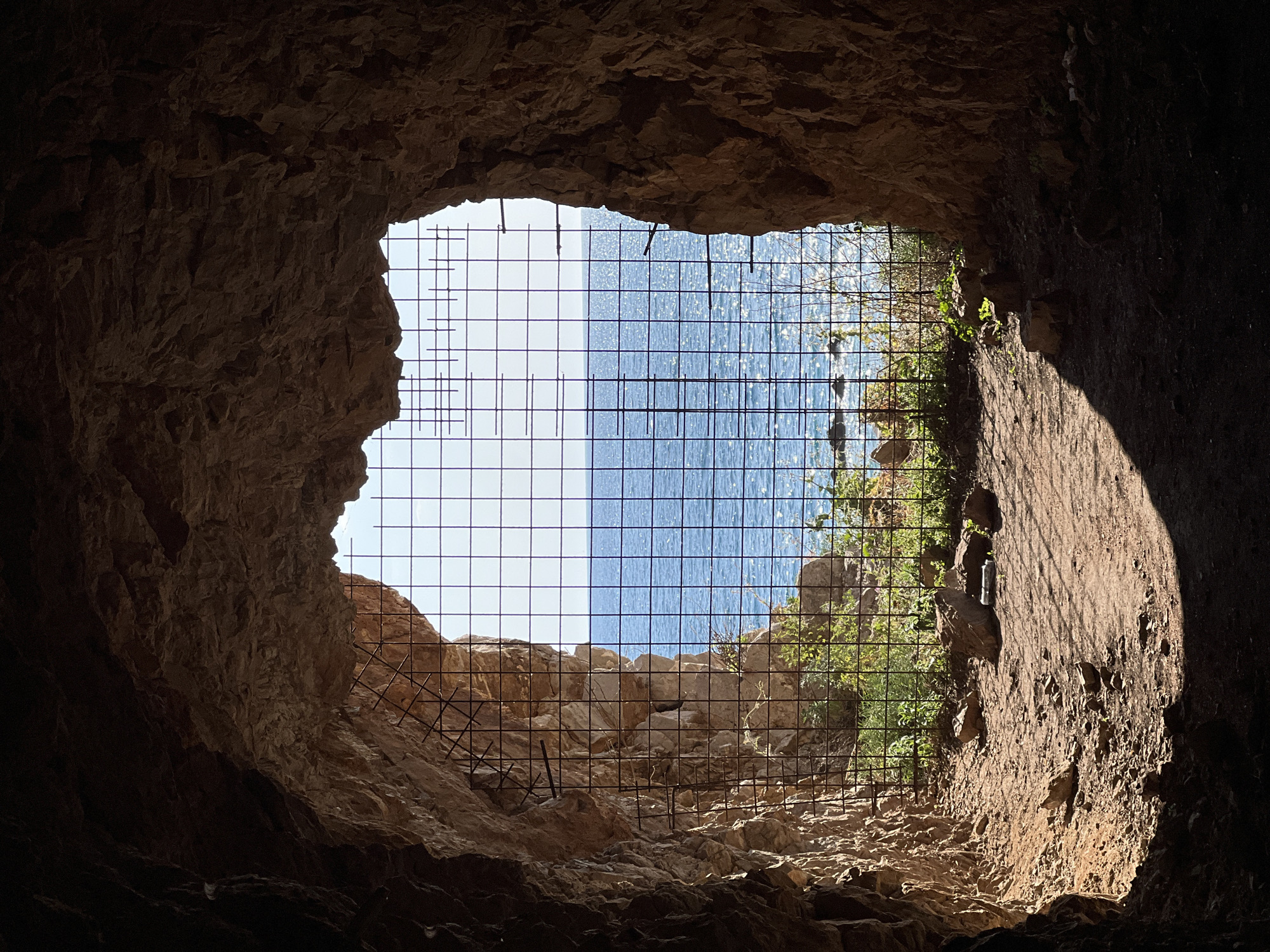 The image shows the view from inside a cave in Perazića Do, Montenegro. The opening of the cave is framed by rocky walls and a rebar grid. Through the grid and cave opening, the Adriatic Sea and coastline are visible. The interior of the cave is dark, with sunlight illuminating sections of the cave wall. The grid obscures some of the view of the sea and coastline.