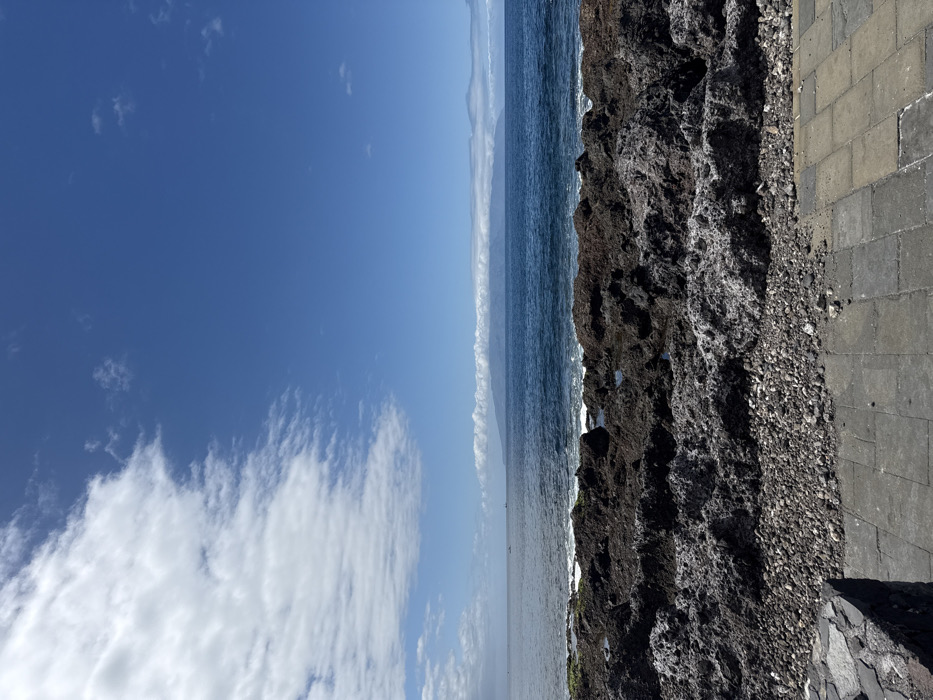 The image captures a coastal scene in Guía de Isora, Spain, presenting a clear sky transitioning from scattered white cumulus clouds on the left to an uninterrupted deep blue on the right. In the immediate foreground, a grey paved walkway with rectangular stones is visible, partially bordered by a low, dark stone structure on the left. This paving transitions into a strip of small, dark grey pebbles and gravel. Beyond the gravel, the shoreline is composed of dark, irregular volcanic rock formations, interspersed with small pools of water and exhibiting patches of green algae. The sea, a dark blue, shows minor surface agitation from waves breaking against the rocks. On the distant horizon, a large landmass identified as an island appears hazy and blue, with its upper slopes obscured by a narrow, horizontal band of white clouds. Two small vessels are visible on the water towards the left of the distant island. No individuals or animate subjects are present in the scene. The lighting suggests full daylight conditions.