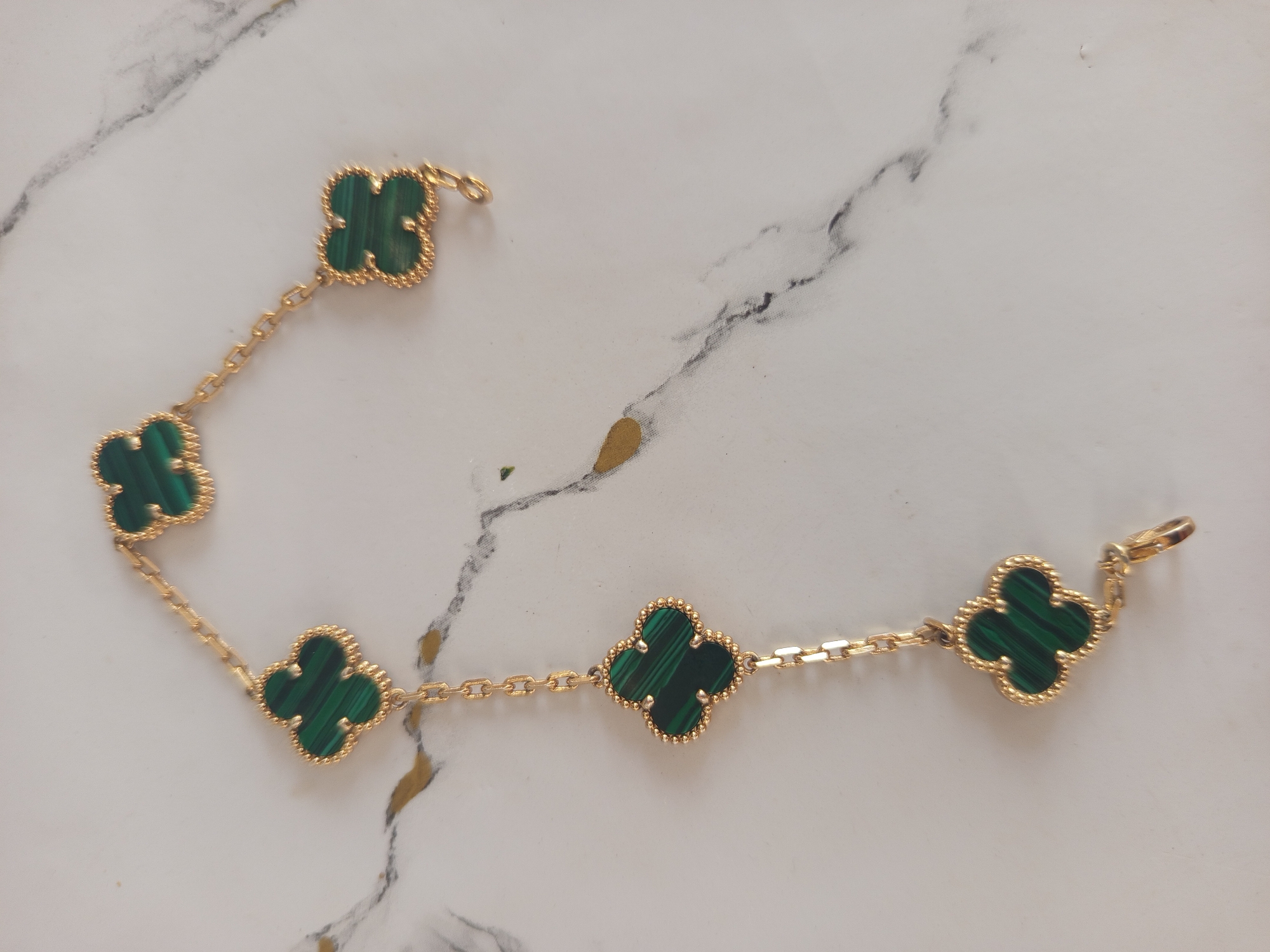 The image shows a gold bracelet with multiple green clover-shaped charms on a white marble surface. The bracelet is laid out diagonally from the top left to the bottom right of the frame. Each charm features a distinctive banded green malachite stone set within a beaded gold border. The chain connecting the charms is delicate and gold. The marble surface has subtle grey veining and a few indistinct gold-colored smudges or markings. There are no people or animals visible in the image. The lighting appears to be even, casting soft shadows. The location of Jardy-Su, Kyrgyzstan is not depicted or relevant to the image content.