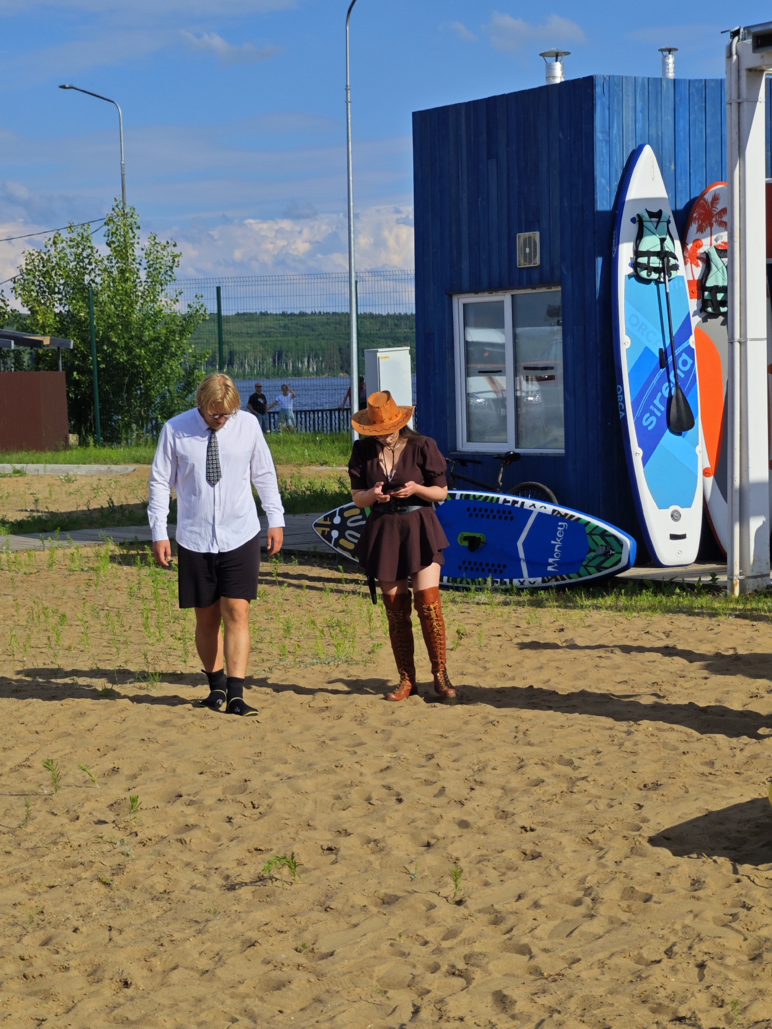 Two individuals are on a sandy beach with a blue wooden building in the background. The person on the left is a man wearing a white dress shirt, black shorts, black socks, and black shoes. He has blonde hair and glasses and is looking down. The person on the right is a woman wearing a brown dress, a brown cowboy hat, and tall brown lace-up boots. She is looking at her phone.
Several paddleboards are leaning against the blue building, some with life vests on them. A bicycle is also visible next to the building. The scene appears to be outdoors on a sunny day with scattered clouds in the sky. In the distance, a body of water and a treeline can be seen. The overall impression is a casual outdoor gathering or activity. There is no visible text in the image.