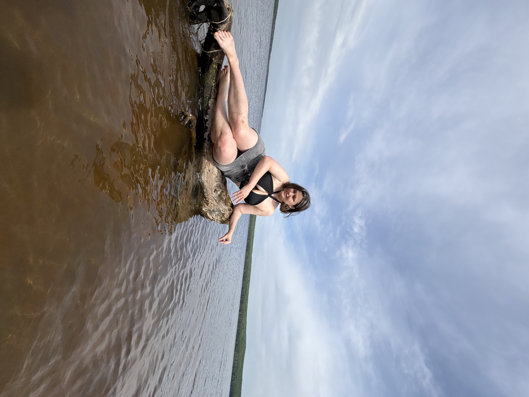 A woman is posing on a log in the water. She is wearing a black bikini top and grey shorts. The water is murky and brown, and the sky is cloudy. There are trees visible on the distant shoreline. The location is implied to be in the city of Chaikovsky, Russia, based on the image title, but this cannot be visually confirmed.