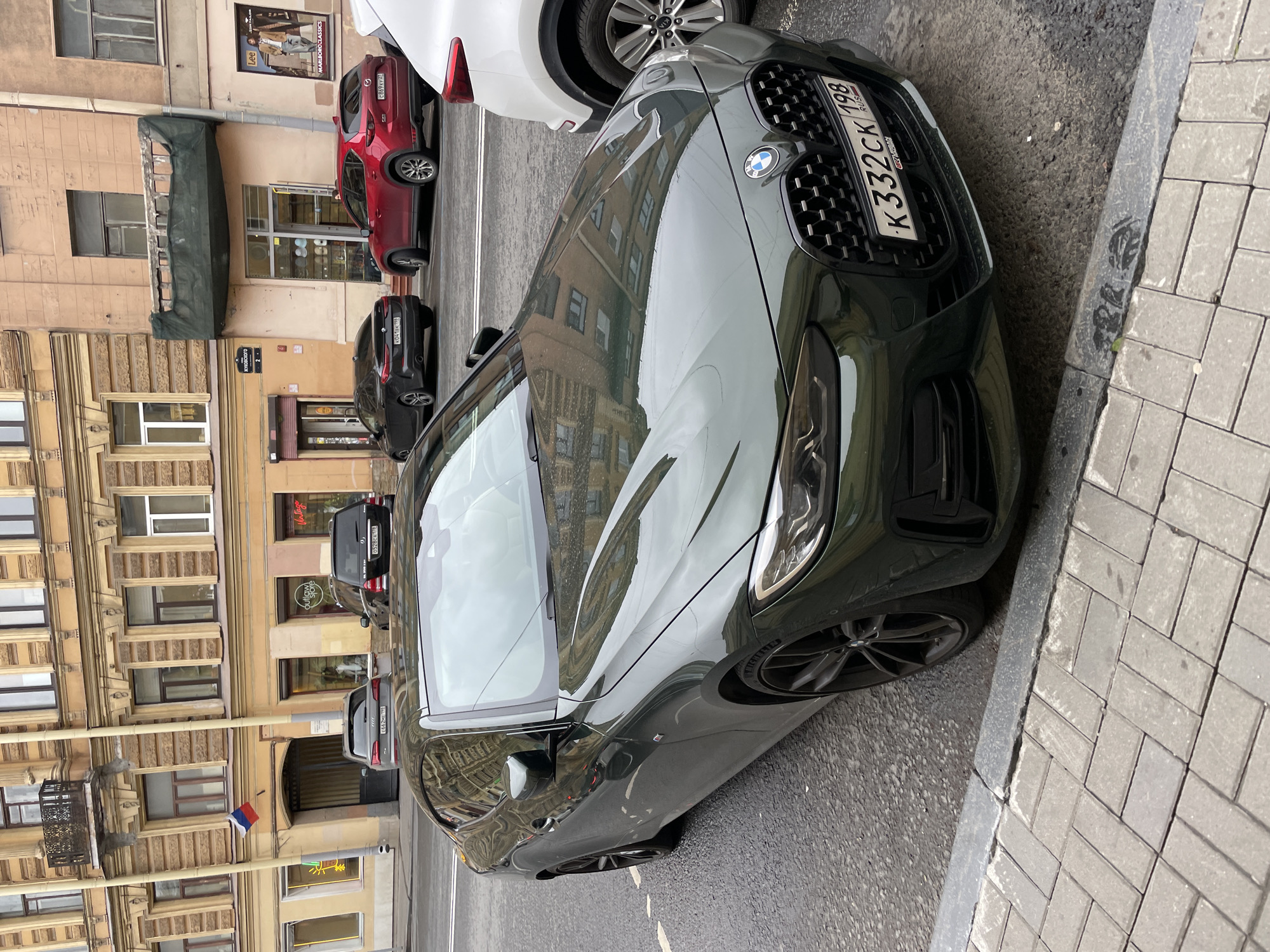 The image shows a dark green BMW car parked on the side of a street in Saint Petersburg, Russia. The car is positioned on the right side of the frame, with its front end facing towards the top. The car's license plate reads "K332CK 198 RUS".
The background of the image features a series of buildings lining the street. Several other cars are parked along the street, including a red Mazda with license plate "C869 KY 22" and a white Kia.
Storefronts of businesses line the bottom of the building facades, with signs visible for "Outlaw store", "Lee", and "MARLBOROCLASSICS". A street sign is visible indicating "OJONOBONAK 2".
The overall impression is a typical urban street scene in Saint Petersburg, with cars parked along the side of the street and buildings lining the background.