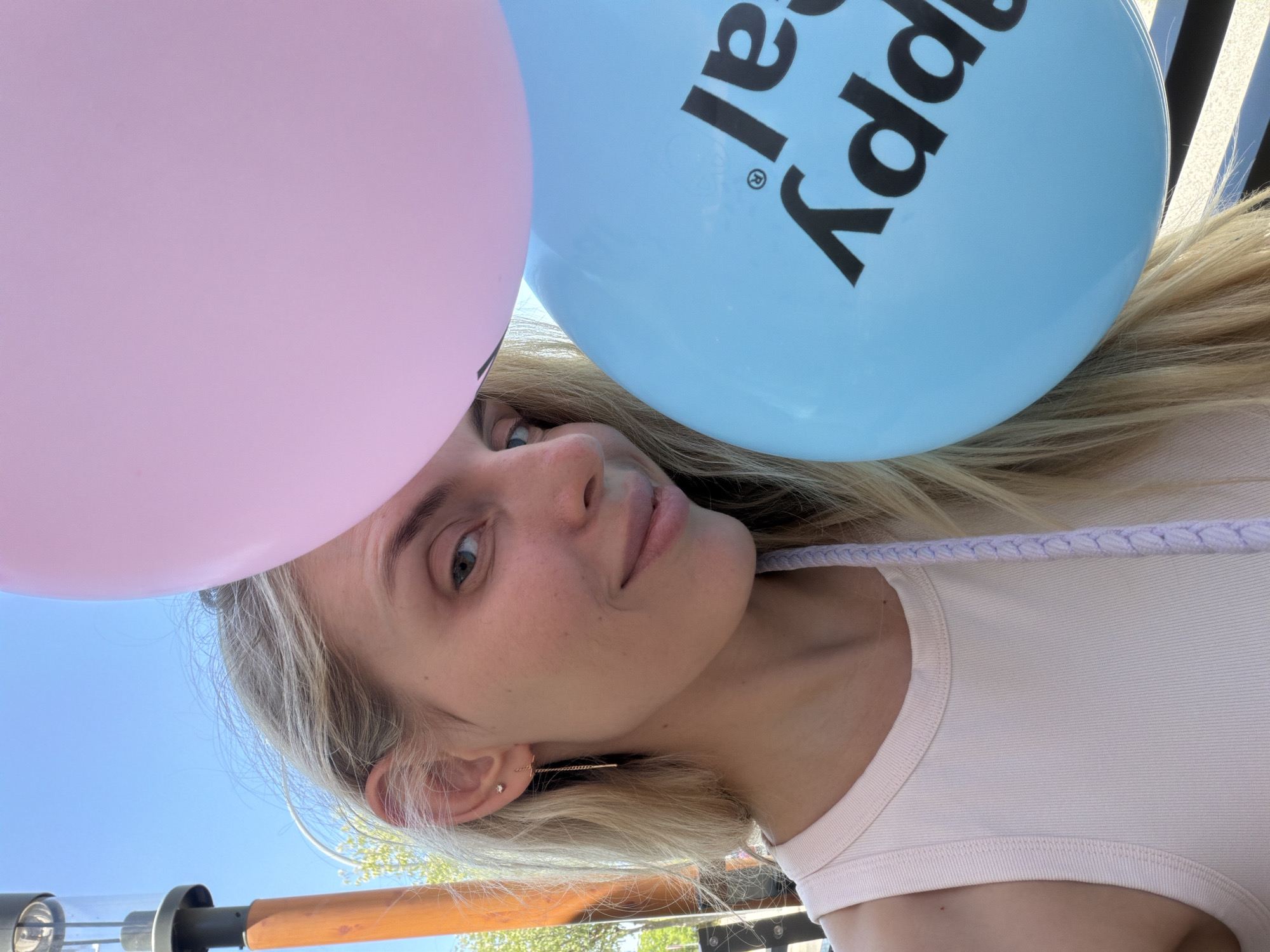 A young woman with blonde hair and blue eyes is partially visible in the frame, holding two balloons. She is wearing a light pink tank top and a delicate gold earring. Her face is turned to the side, and she has a slight smile.
One balloon is pink, and the other is light blue with black lettering. The blue balloon has the partial text "appy" and "al" visible, suggesting it says "Happy Birthday". The image appears to be taken outdoors on a sunny day, as indicated by the bright lighting and the clear blue sky in the background. There are some trees and structures in the background, which are blurred. The overall mood is cheerful and celebratory. The woman's hair is flowing, and she appears to be looking up. The scene is shot from a low angle, with the balloons dominating the upper portion of the image.