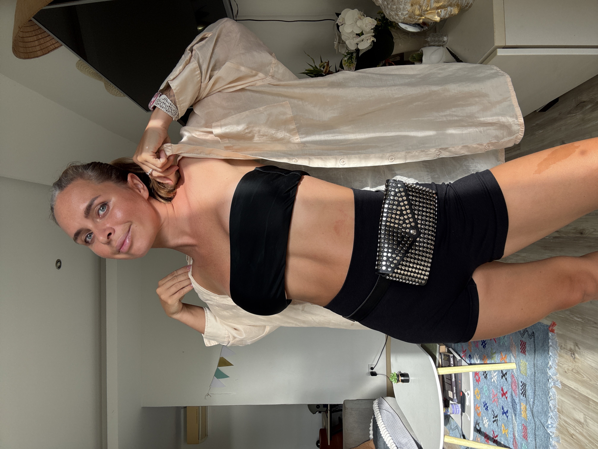 In a room on the island of Ko Samui, Thailand, a woman stands in front of a white wall, bathed in natural sunlight. She is wearing a black strapless top, black shorts, and an oversized, light-colored button-up shirt worn open over her shoulders. A studded black fanny pack is cinched at her waist. Her hair is pulled back in a ponytail. The background shows a living area with a sofa, a coffee table with a plant on it, and a rug. To the right, a white cabinet holds decorative items, including a white orchid and a large, gold-leafed Buddha head bust.
